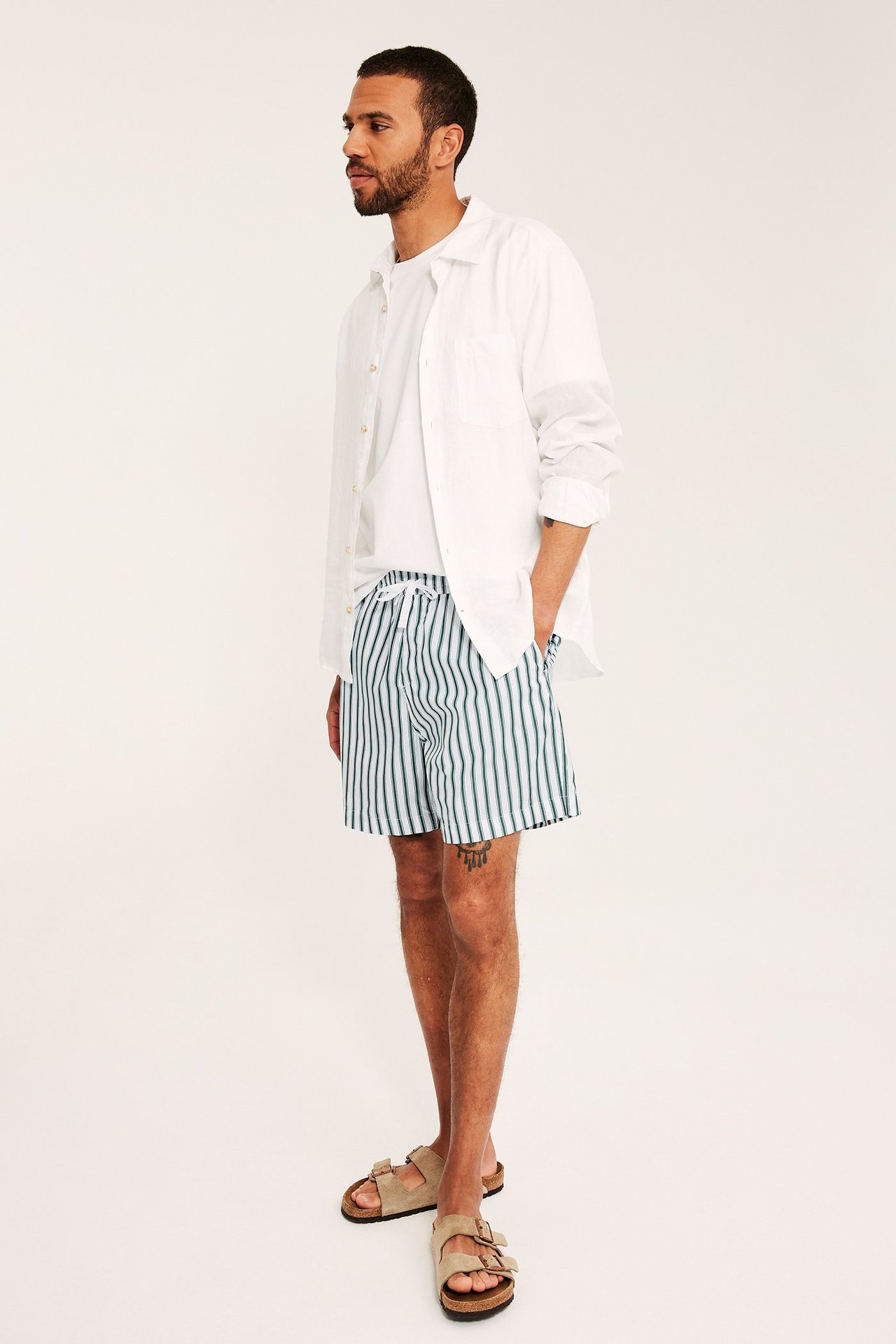 Mężczyzna w biało-zielonych bawełnianych szortach w paski, białym t-shircie, otwartej białej koszuli i beżowych sandałach. Widok z boku.