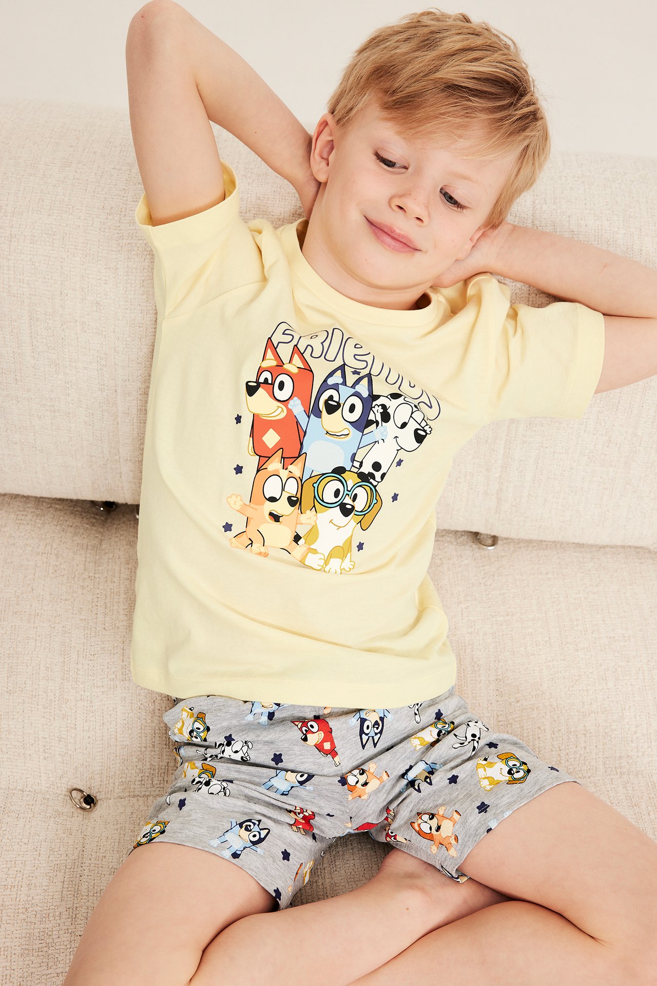 Barn pojke i ljusgul pyjamas med Bluey-tryck, sittande på soffa, avslappnad pose.