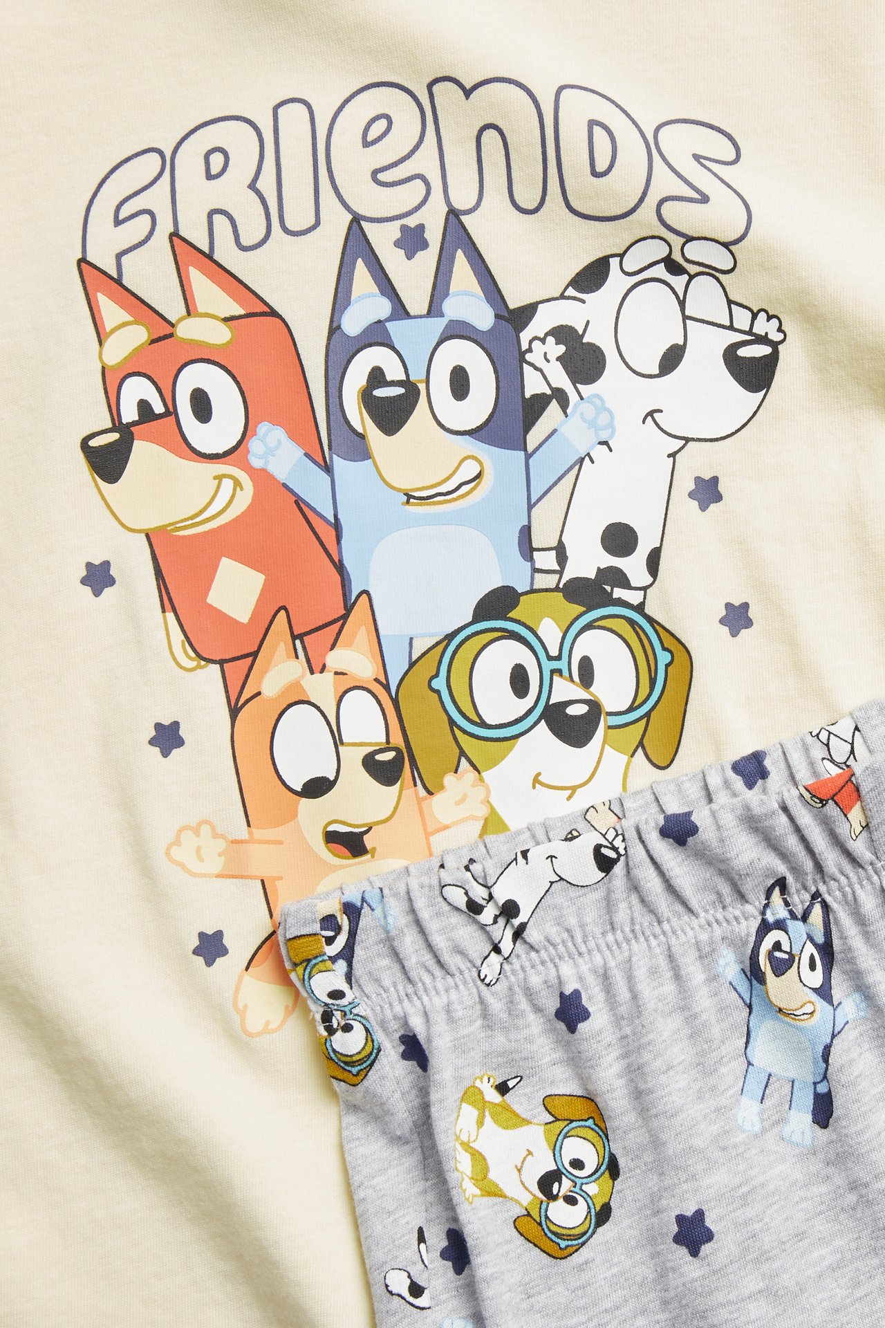 Barns ljusgula pyjamastopp med Bluey-tryck och gråmönstrade byxor, närbild framifrån.