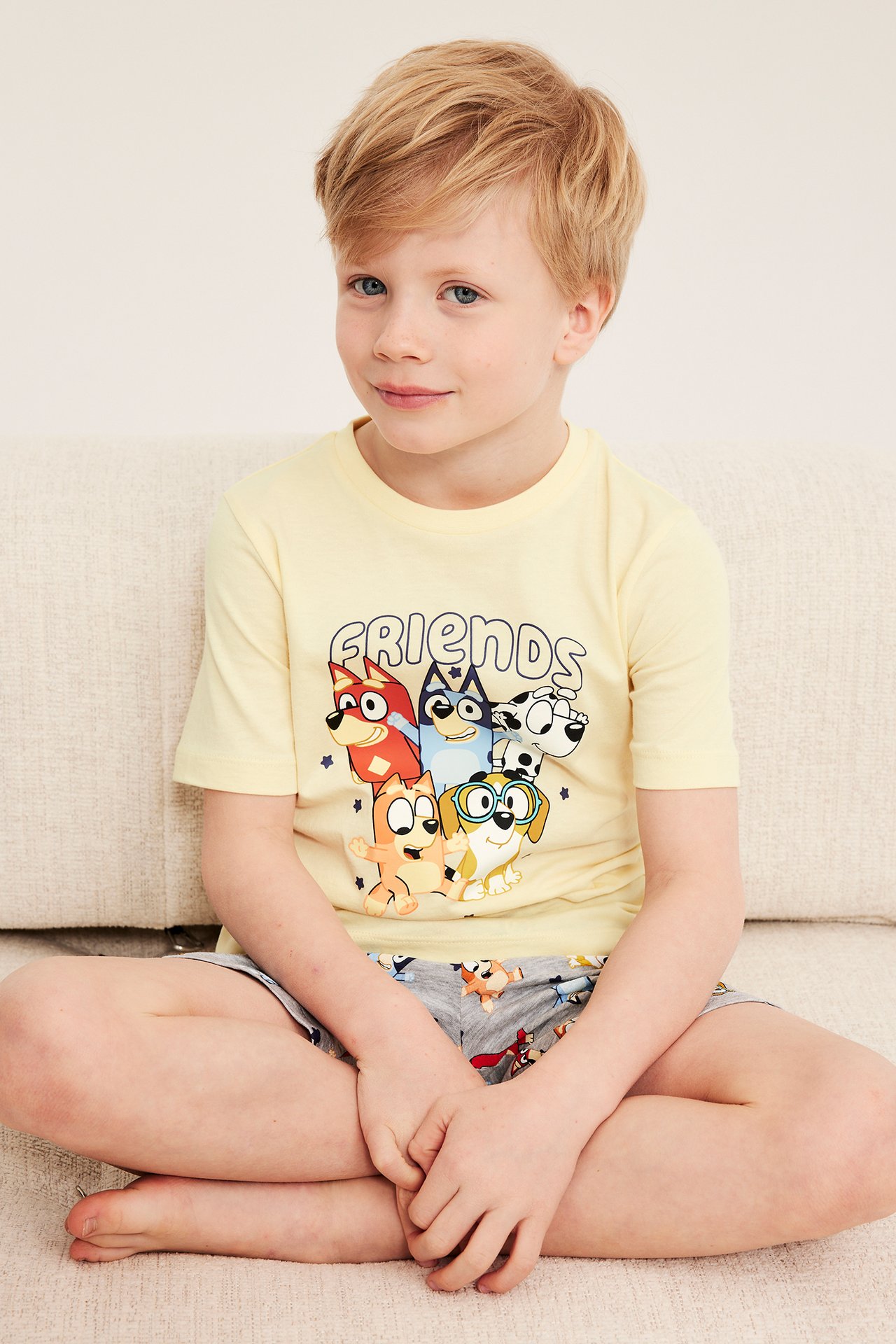 Frontvy av ett barn i ljusgul pyjamaströja i bomull med Bluey-tryck och gråmönstrade shorts.