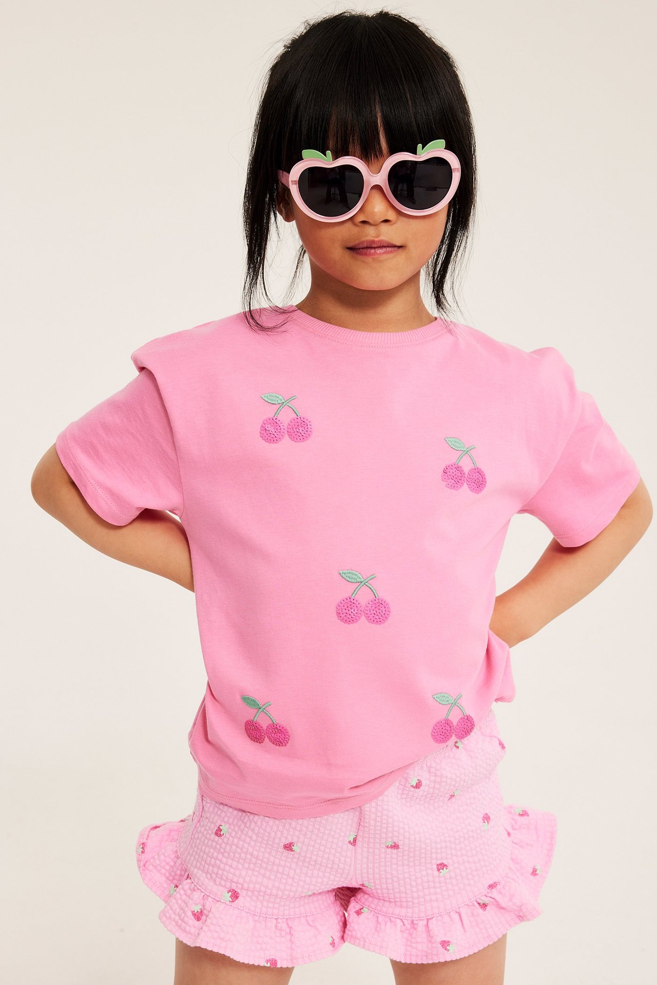 Kid har på seg lys rosa jordbær-shorts med volanger, rosa kirsebær-t-skjorte og rosa hjerte-solbriller.