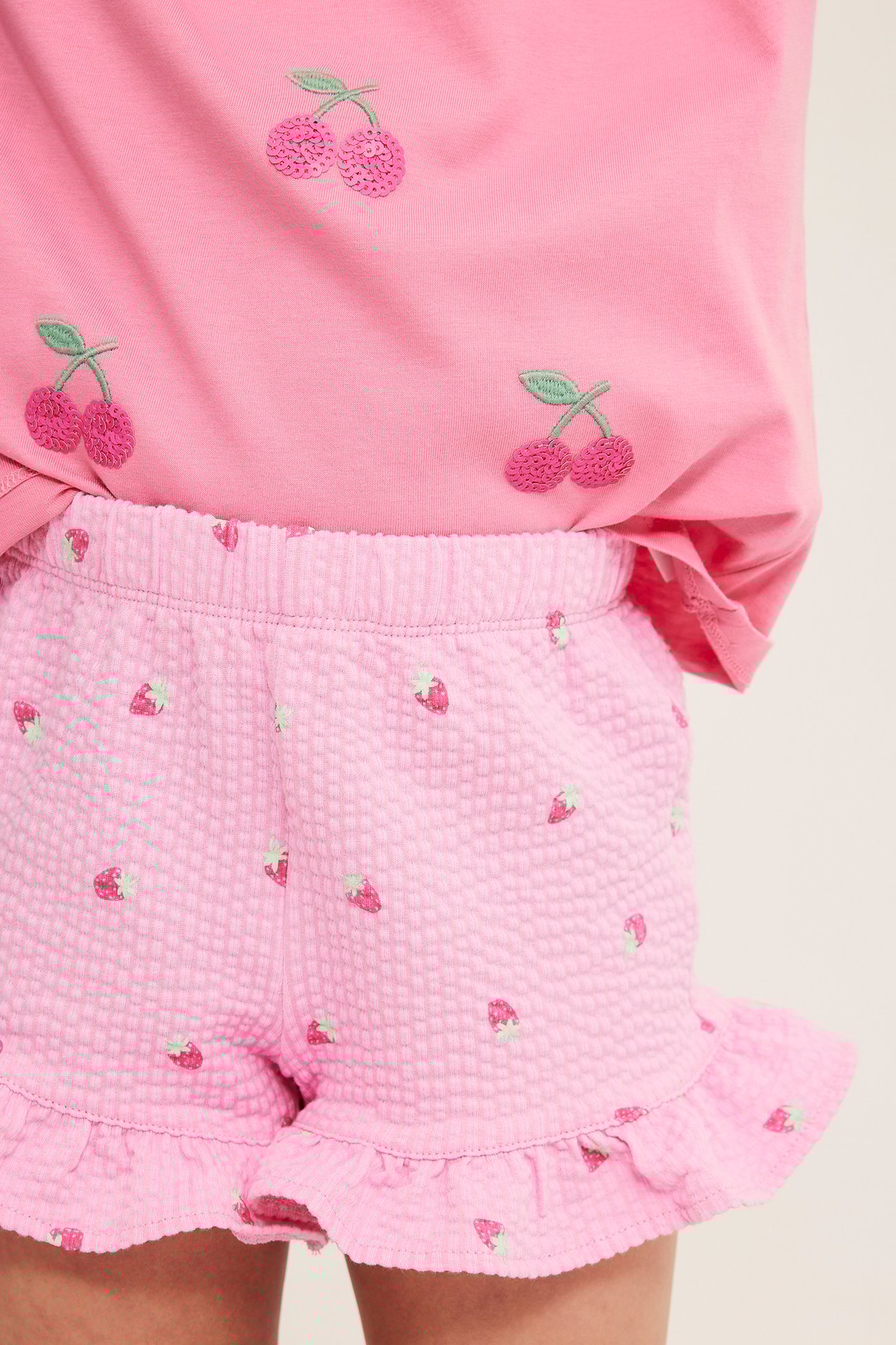 Närbild: Barns ljusrosa jordgubbsshorts med volanger, samt en rosa körsbärs-t-shirt.