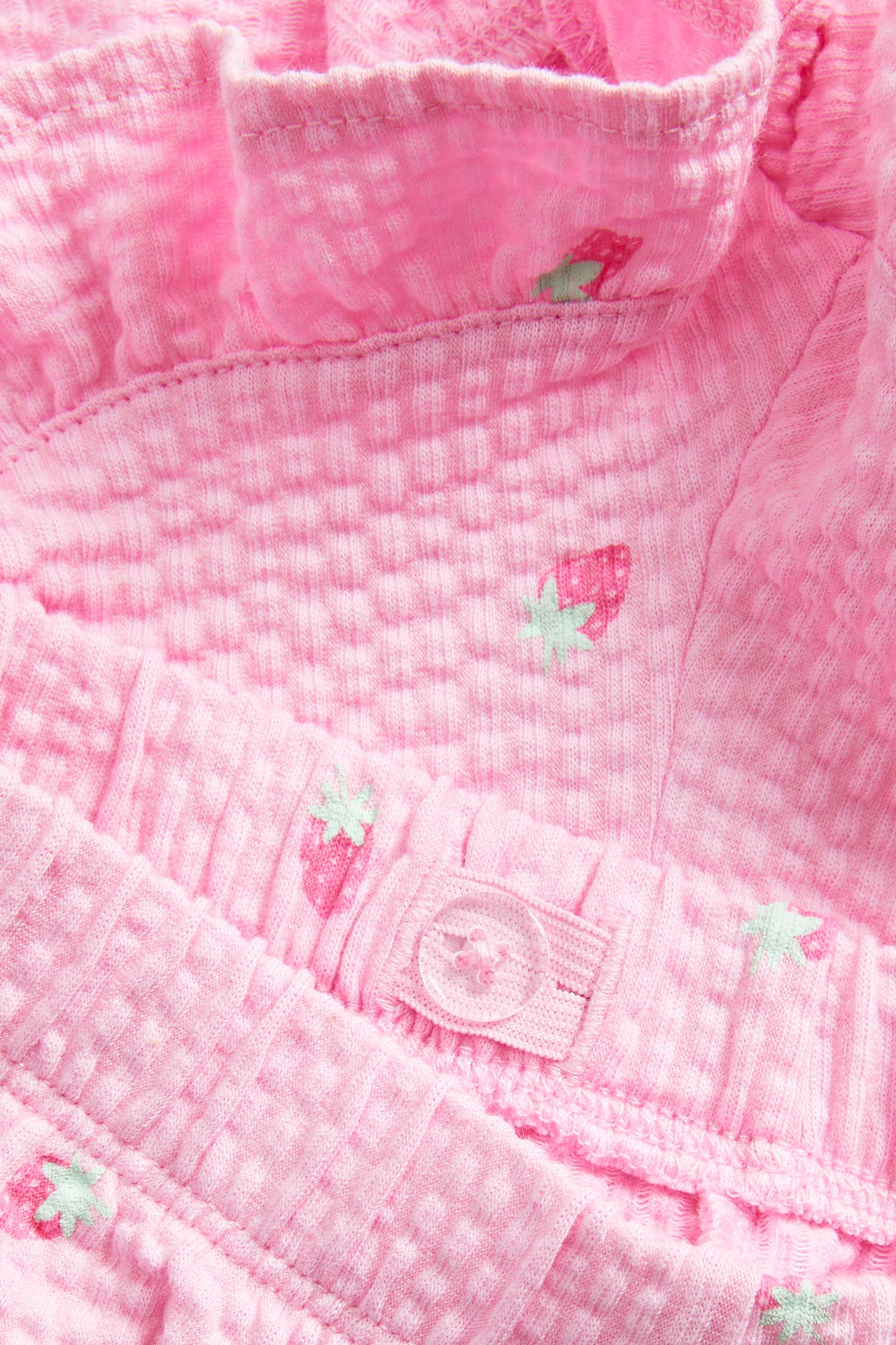 Lähikuva vaaleanpunaisista strukturoiduista puuvillashortseista mansikkakuviolla, röyhelöreunalla ja säädettävällä vyötärönapilla.