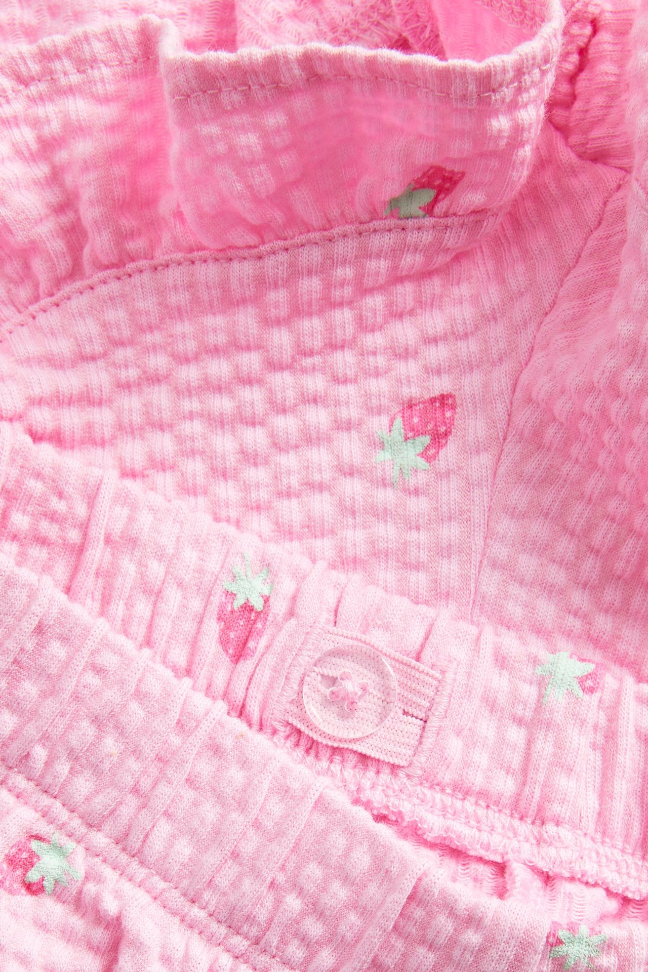 Zbliżenie na jasnoróżowe, strukturyzowane bawełniane szorty w truskawkowy wzór z falbanką i regulowanym guzikiem w pasie.