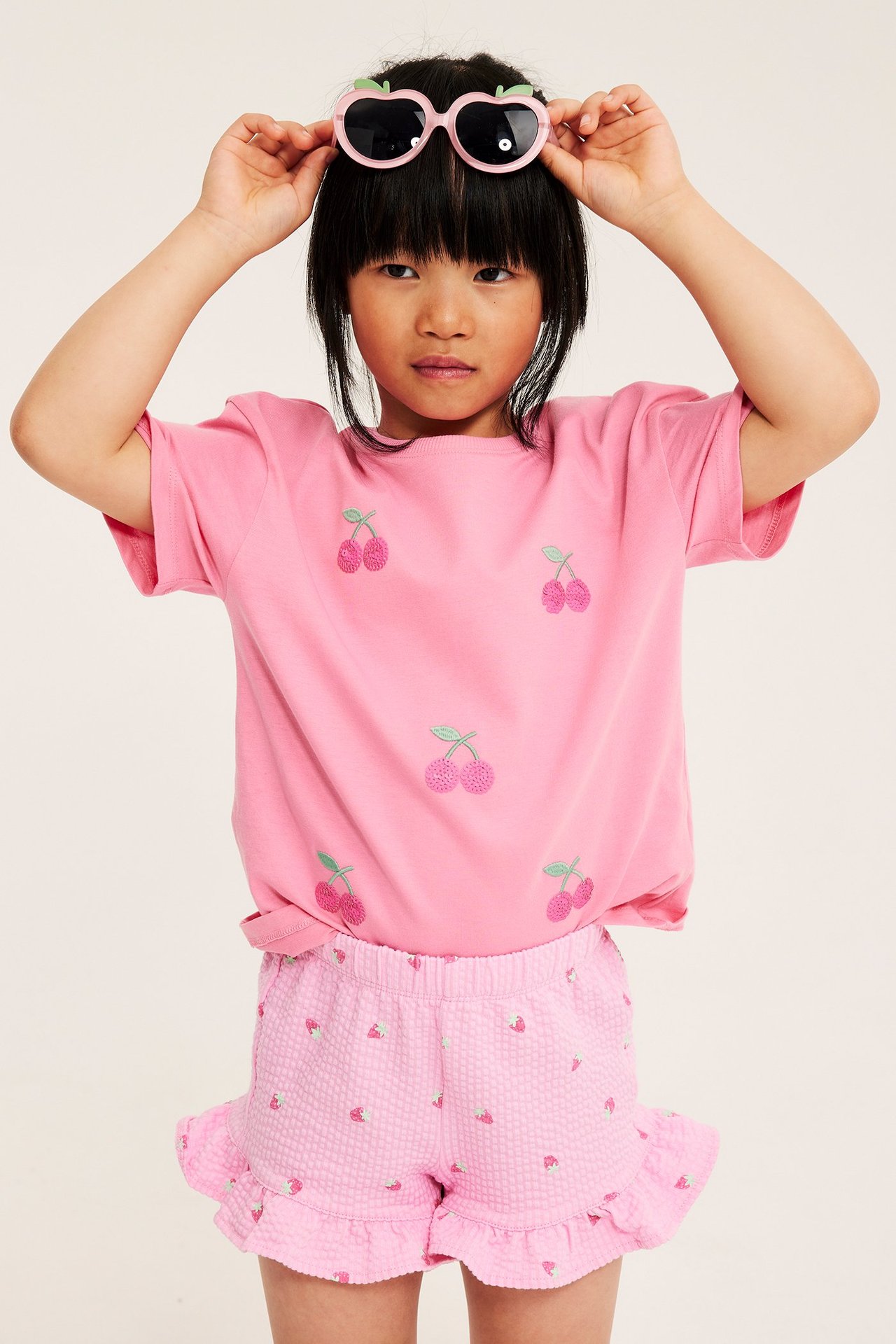 Et barn har på seg lys rosa jordbærmønstrede shorts med volanger, en rosa kirsebær-t-skjorte og kirsebær-solbriller.