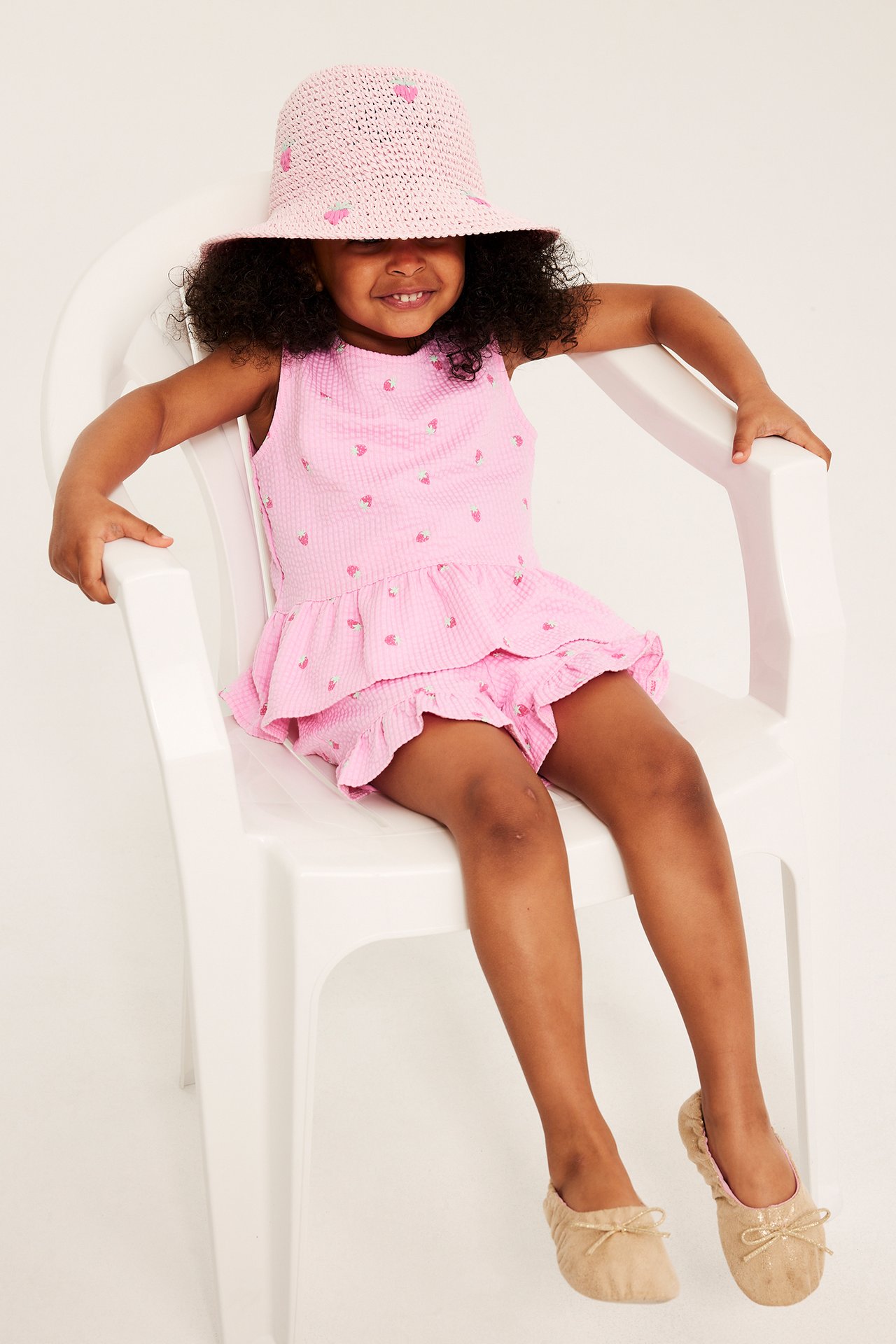 Framifrån: Barn i ljusrosa jordgubbsmönstrad peplumtopp, matchande shorts, rosa hatt och beiga ballerinaskor.