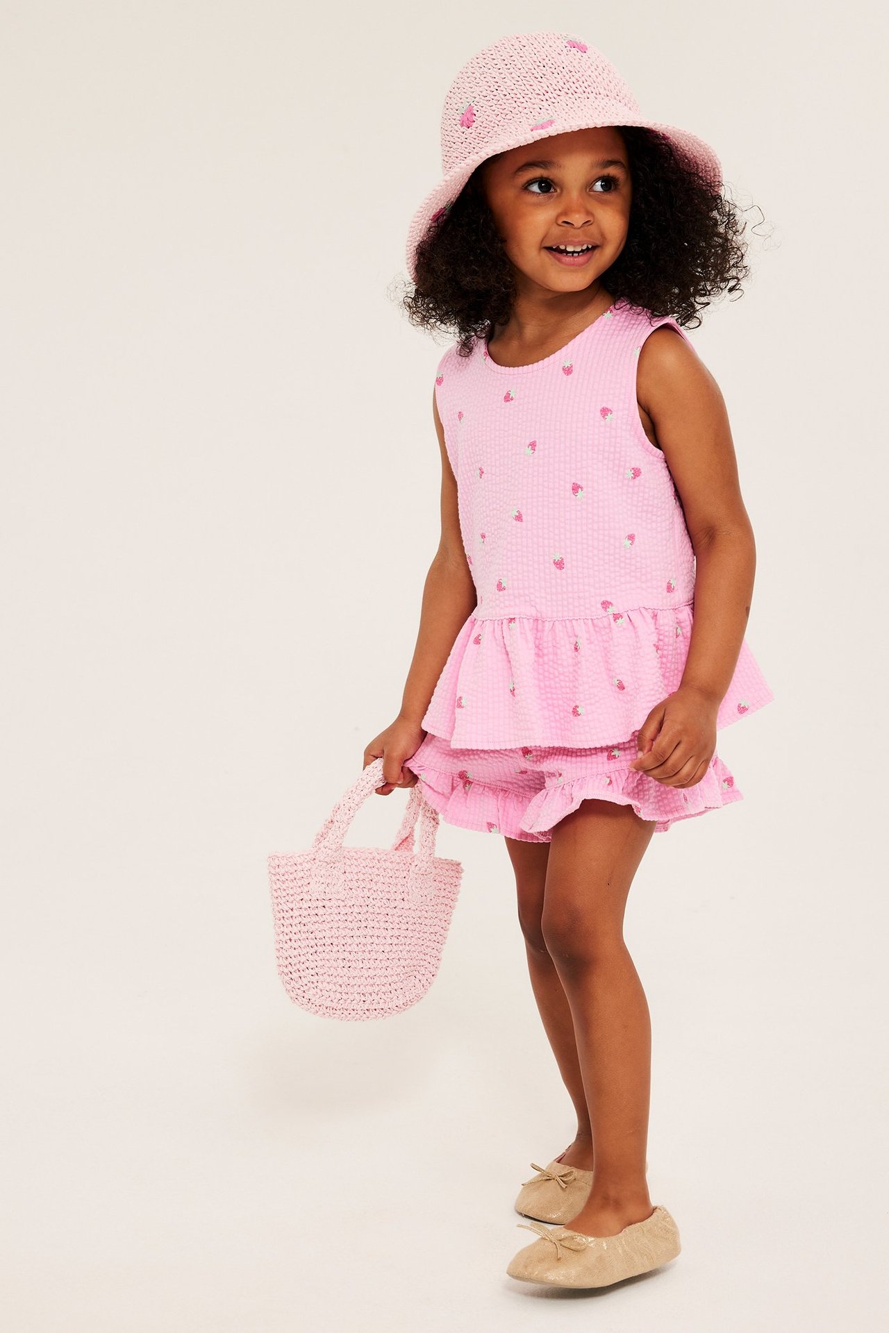 Edestäpäin: Lapsi mansikkakuvioisessa vaaleanpunaisessa seersucker-peplumtopissa, shortsit, hattu, laukku.