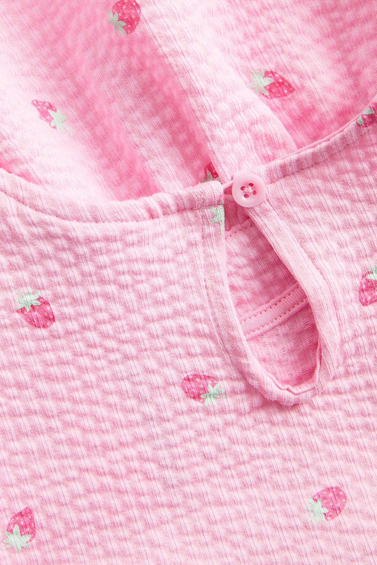 Lähikuva: lasten vaaleanpunainen, strukturoitu peplumtoppi mansikkakuviolla, nappikiinnitys takana.