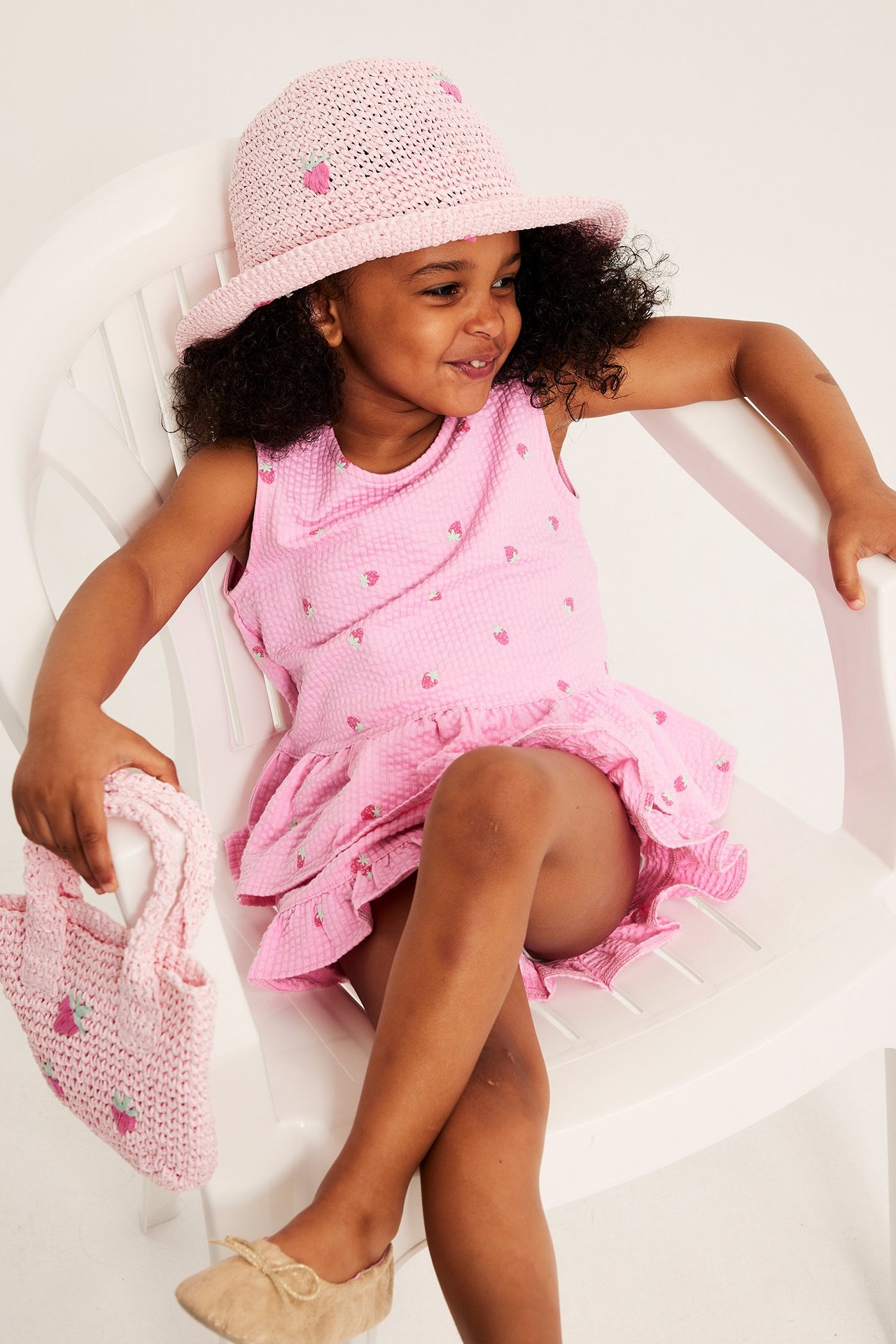 Widok z przodu dziecka w różowym topie peplum we wzór truskawek, z pasującym kapeluszem i torbą.
