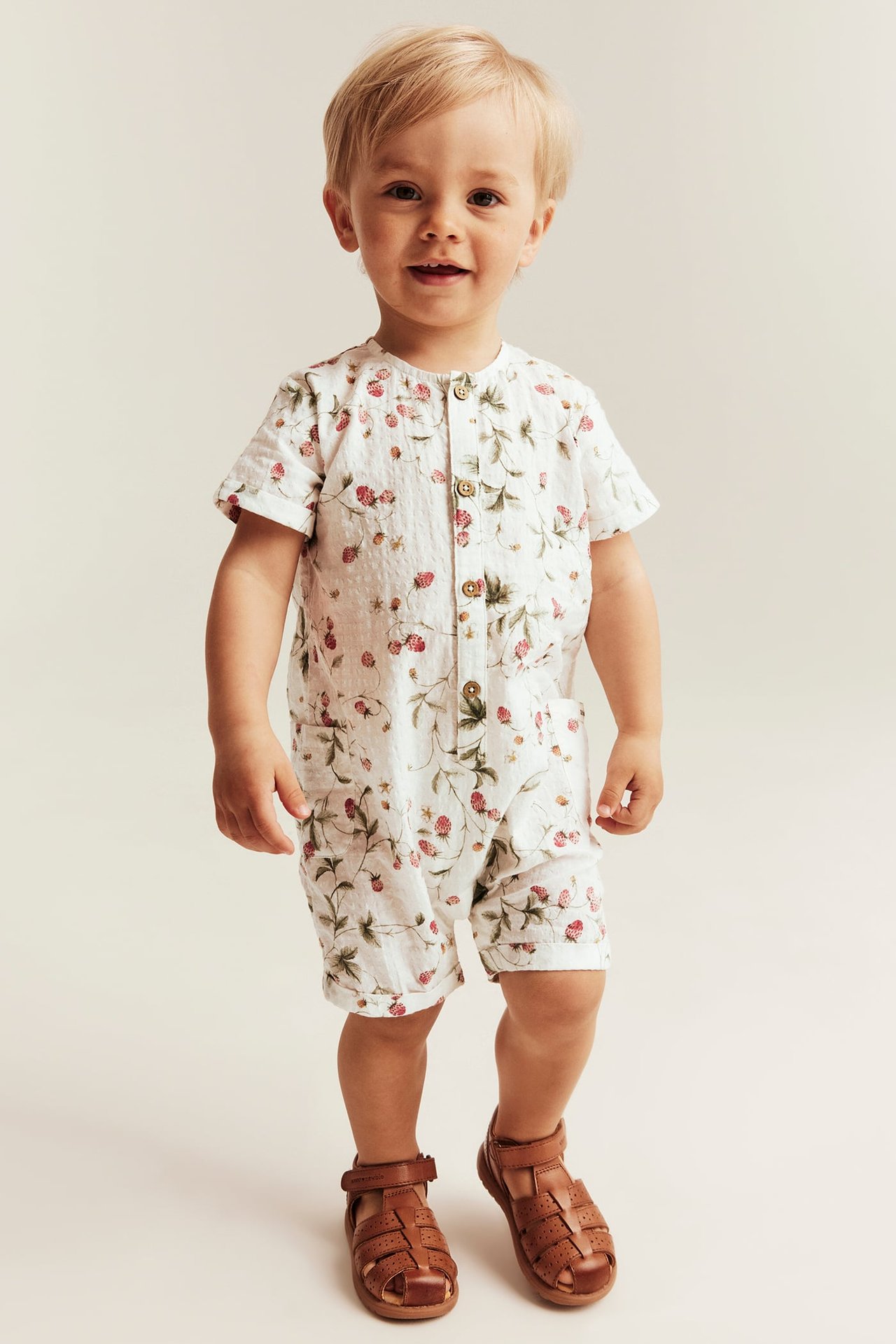 Framifrån, ett baby bär en offwhite bomullsromper med smultronmönster och bruna sandaler.