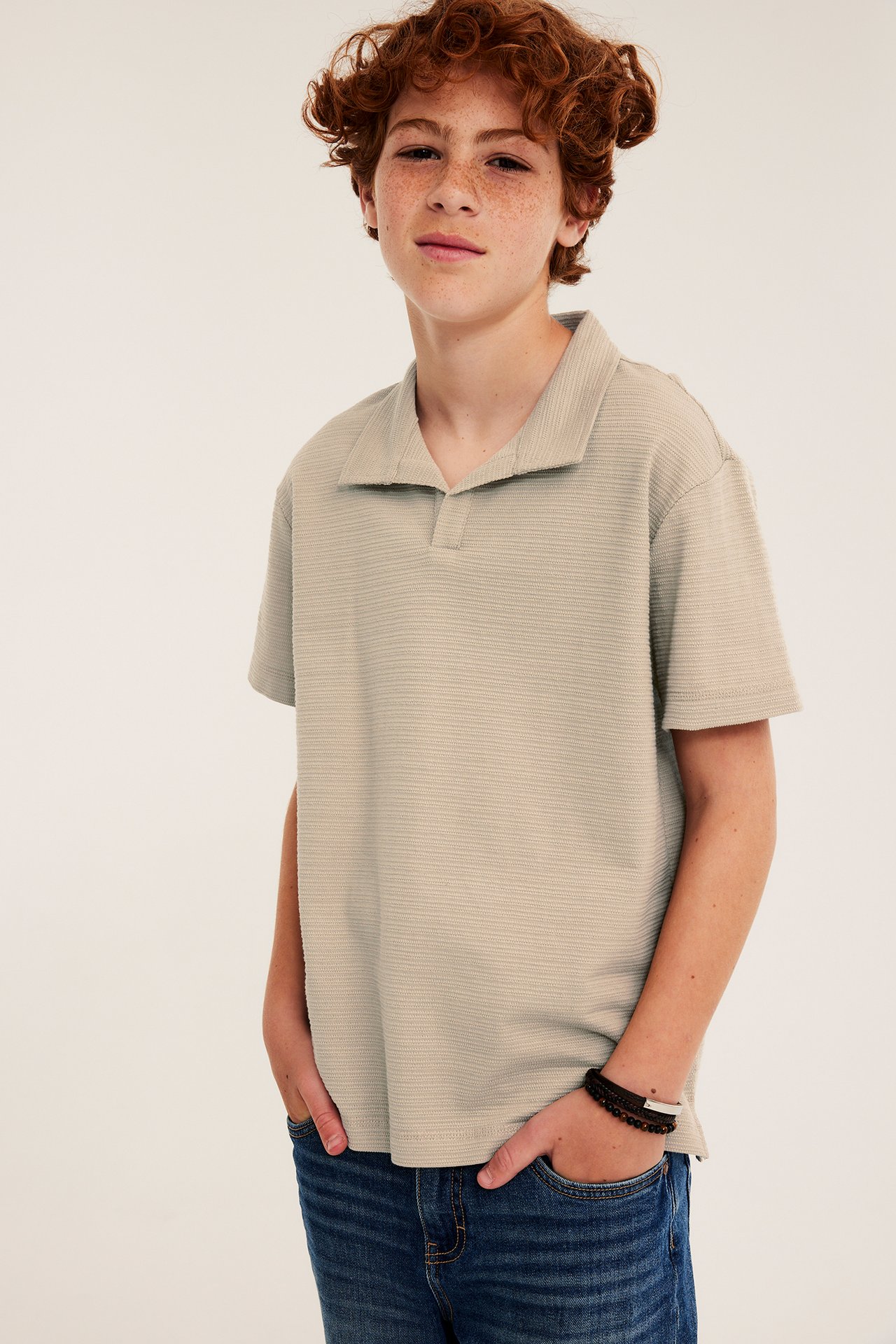 Framifrån: kid i beige strukturerad pikétröja, blåa jeans och svarta samt bruna armband.