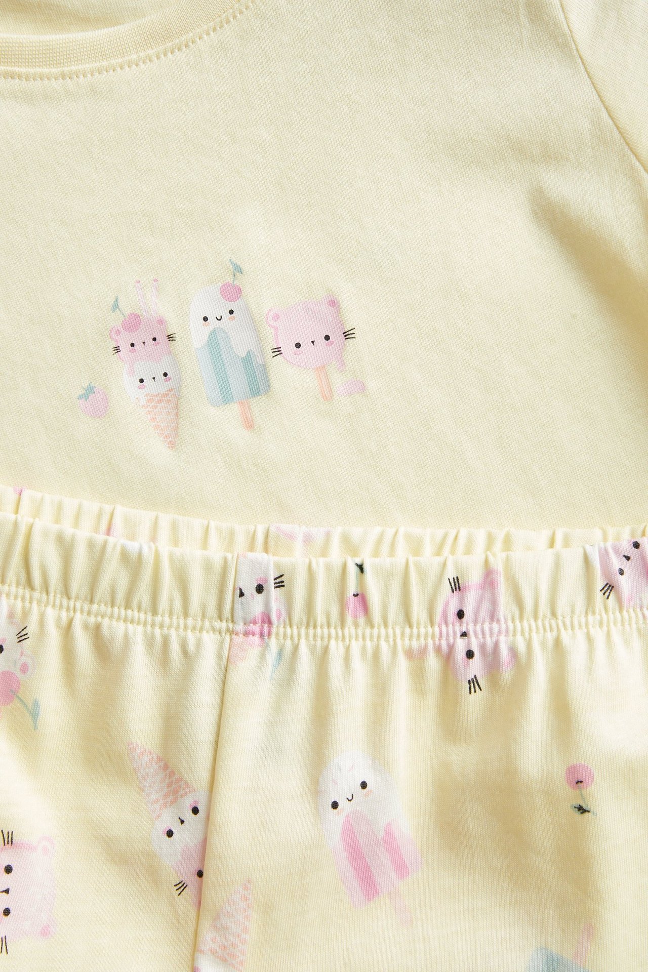 Zbliżenie na jasnożółtą, dziecięcą bawełnianą piżamę z krótkim rękawem, z kolorowym nadrukiem lodów i elastycznym pasem.