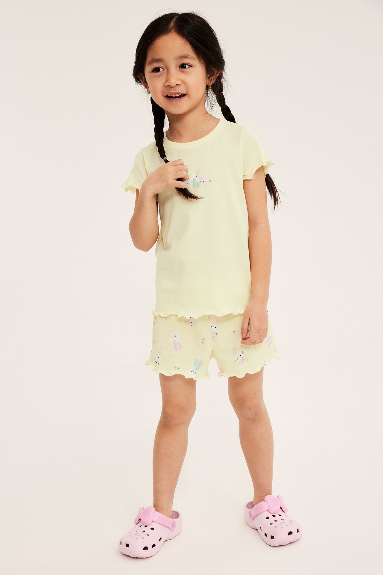 Widok z przodu: Dziecko w jasnożółtej bawełnianej piżamie z falbanami, lodowym nadrukiem i różowymi klapkami.
