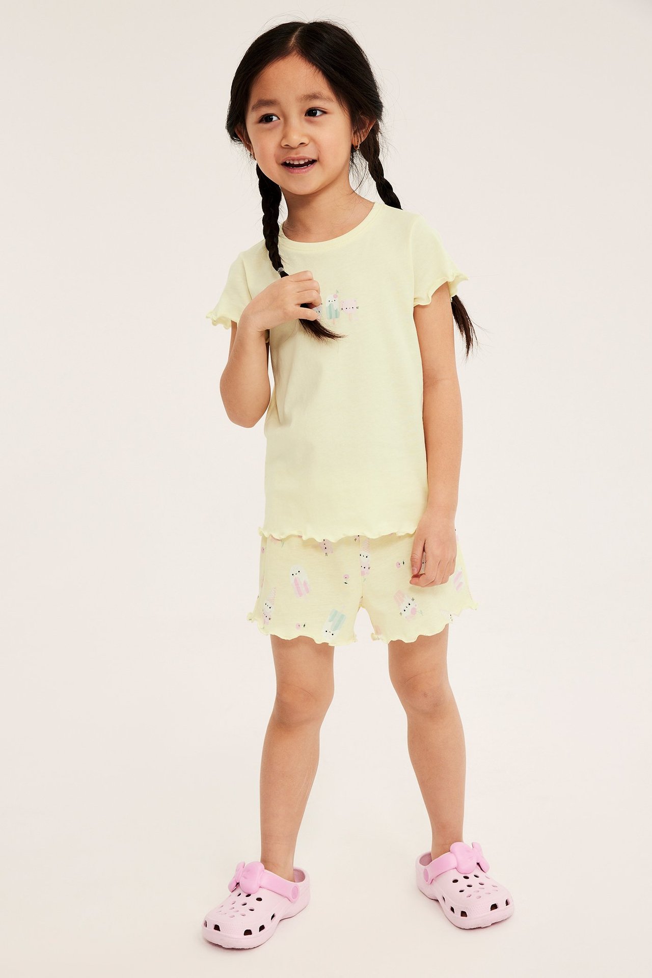 Widok z przodu: Dziecko w jasnożółtej bawełnianej piżamie z falbanami, lodowym nadrukiem i różowymi klapkami.