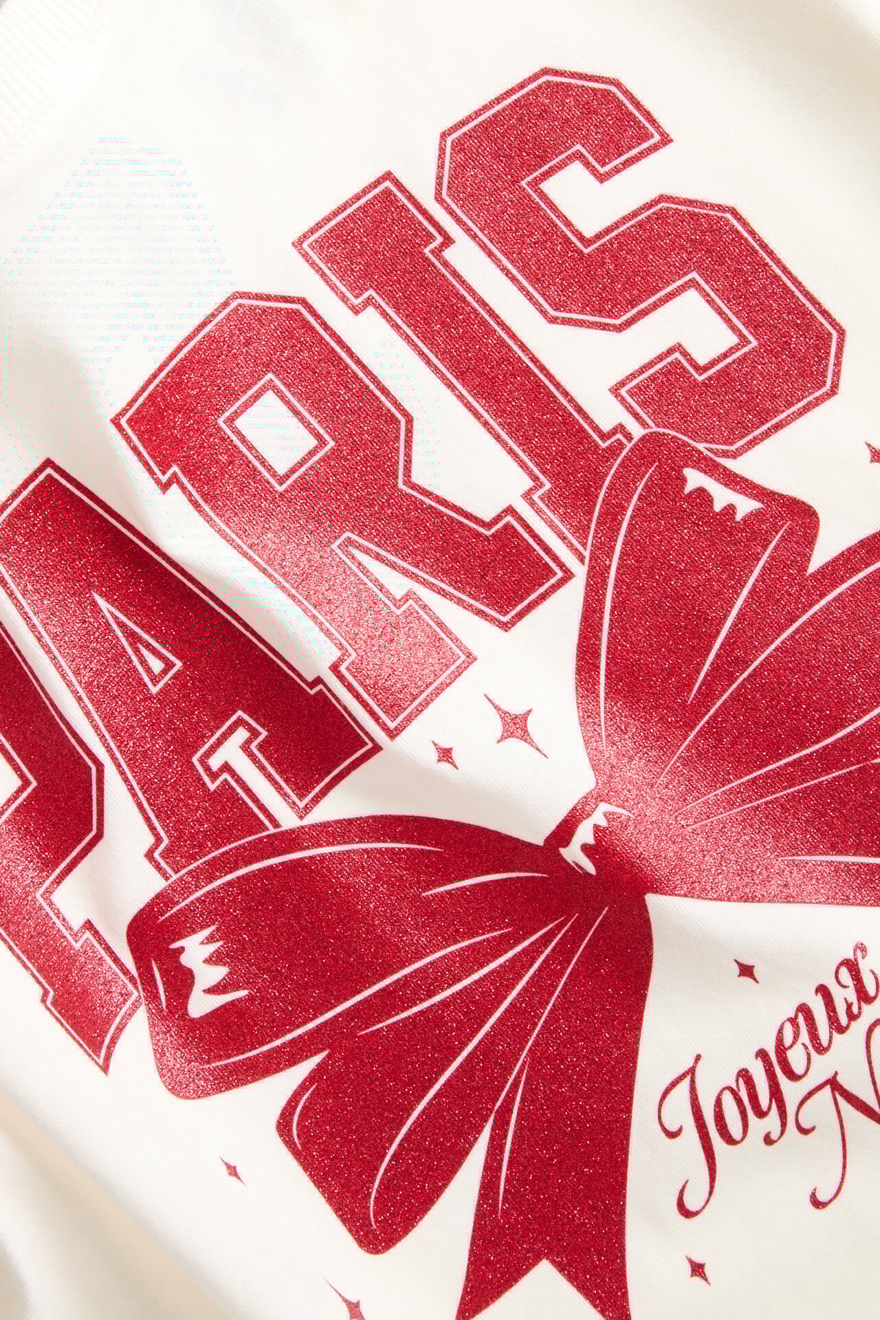 Närbild av en vit bomulls-t-shirt med rött tryck: "PARIS", en rosett och "Joyeux N" text.