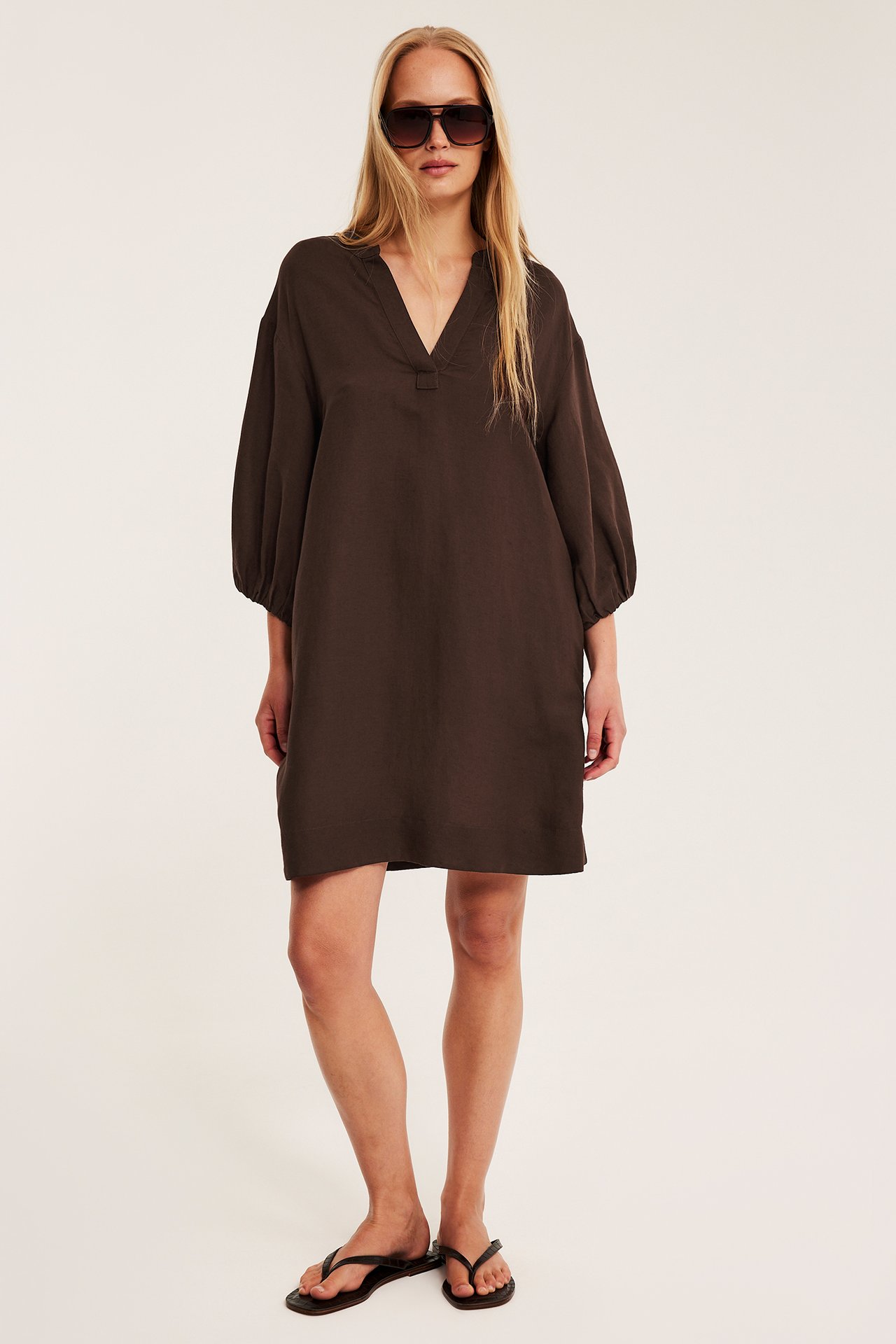 Kvinna framifrån i mörkbrun V-ringad klänning i linneblandning med ballongärmar, mörka solglasögon och svarta flip-flops.