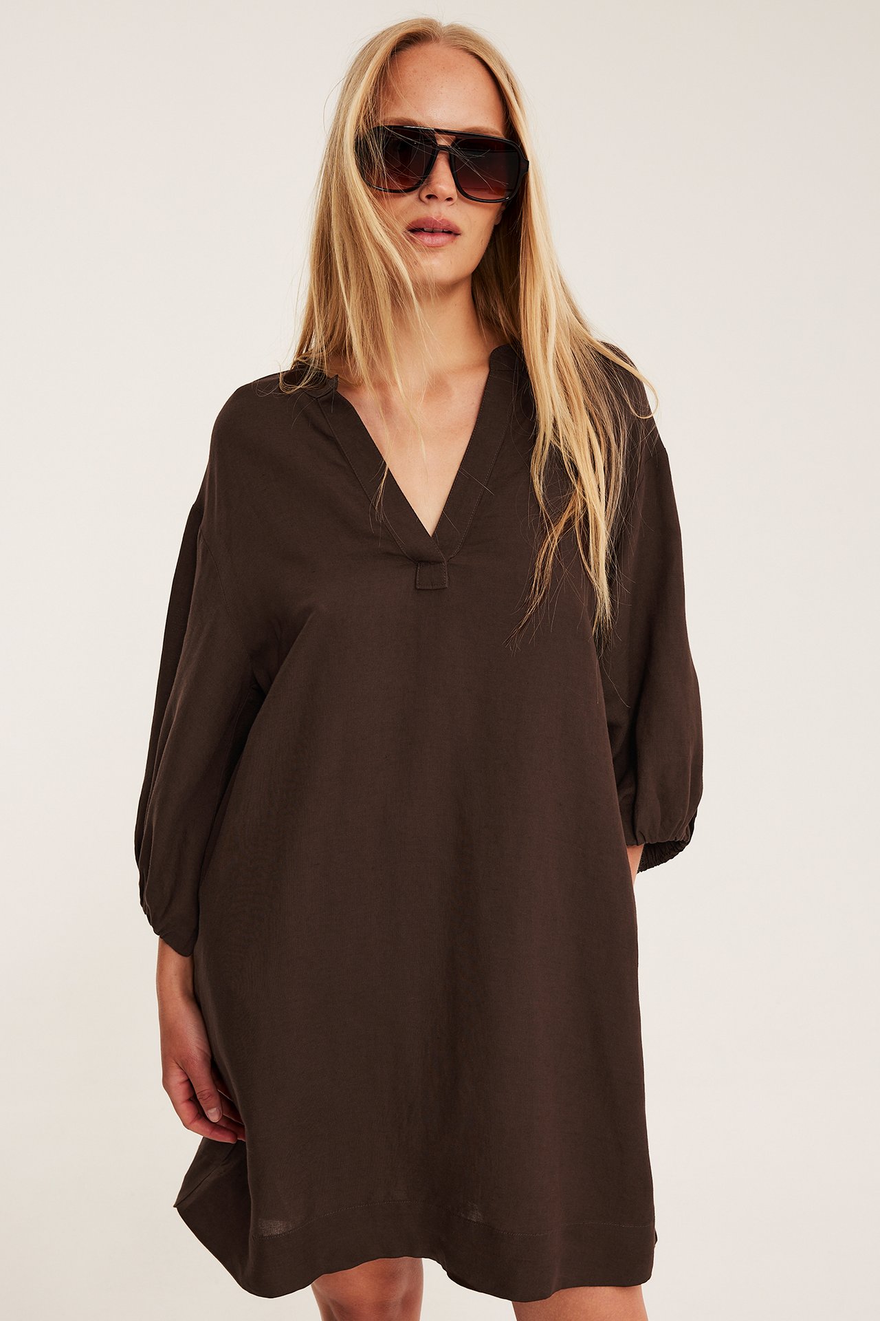 Widok z przodu kobiety w ciemnobrązowej sukience z dekoltem V, z mieszanki lnu, bufiastymi rękawami i ciemnymi okularami.