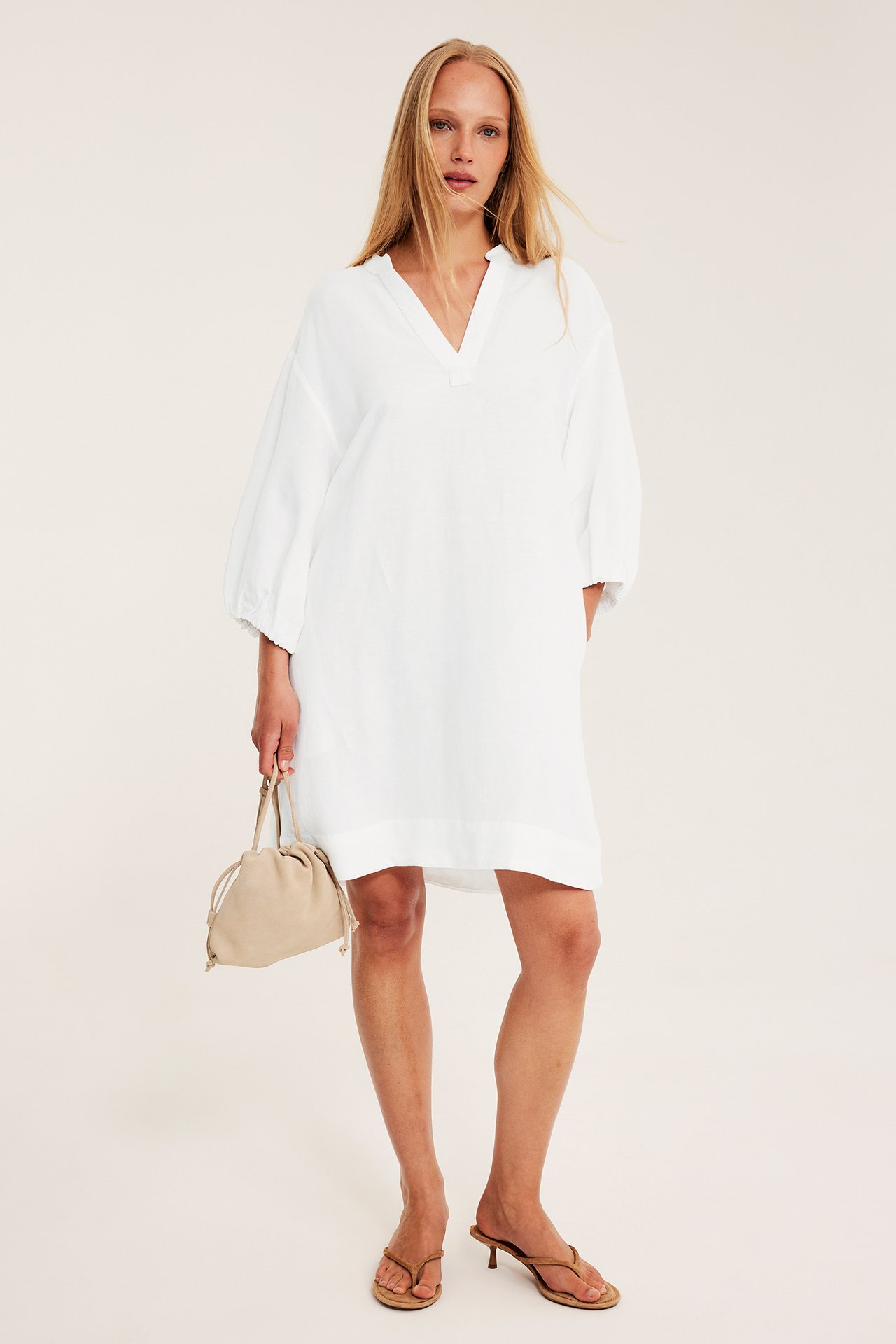 Kvinna i vit V-ringad linneklänning med ballongärm, håller beige dragskoväska, bär bruna klacksandaler.
