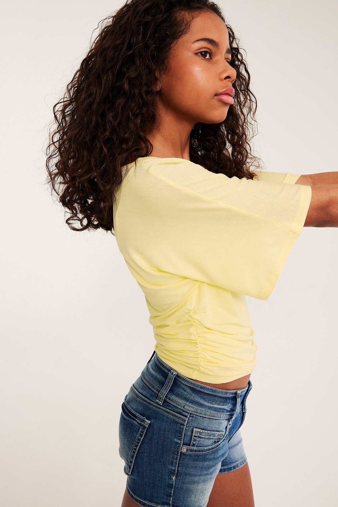 Boczny widok: dziewczynka w jasnożółtym topie motylkowym z marszczoną talią i niebieskimi szortami.