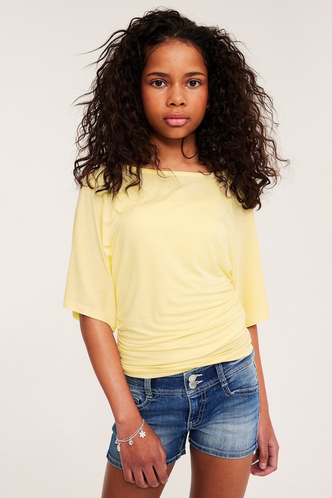Dziecko nosi jasnożółty top z drapowaniem i marszczeniami, niebieskie szorty jeansowe oraz srebrną bransoletkę.