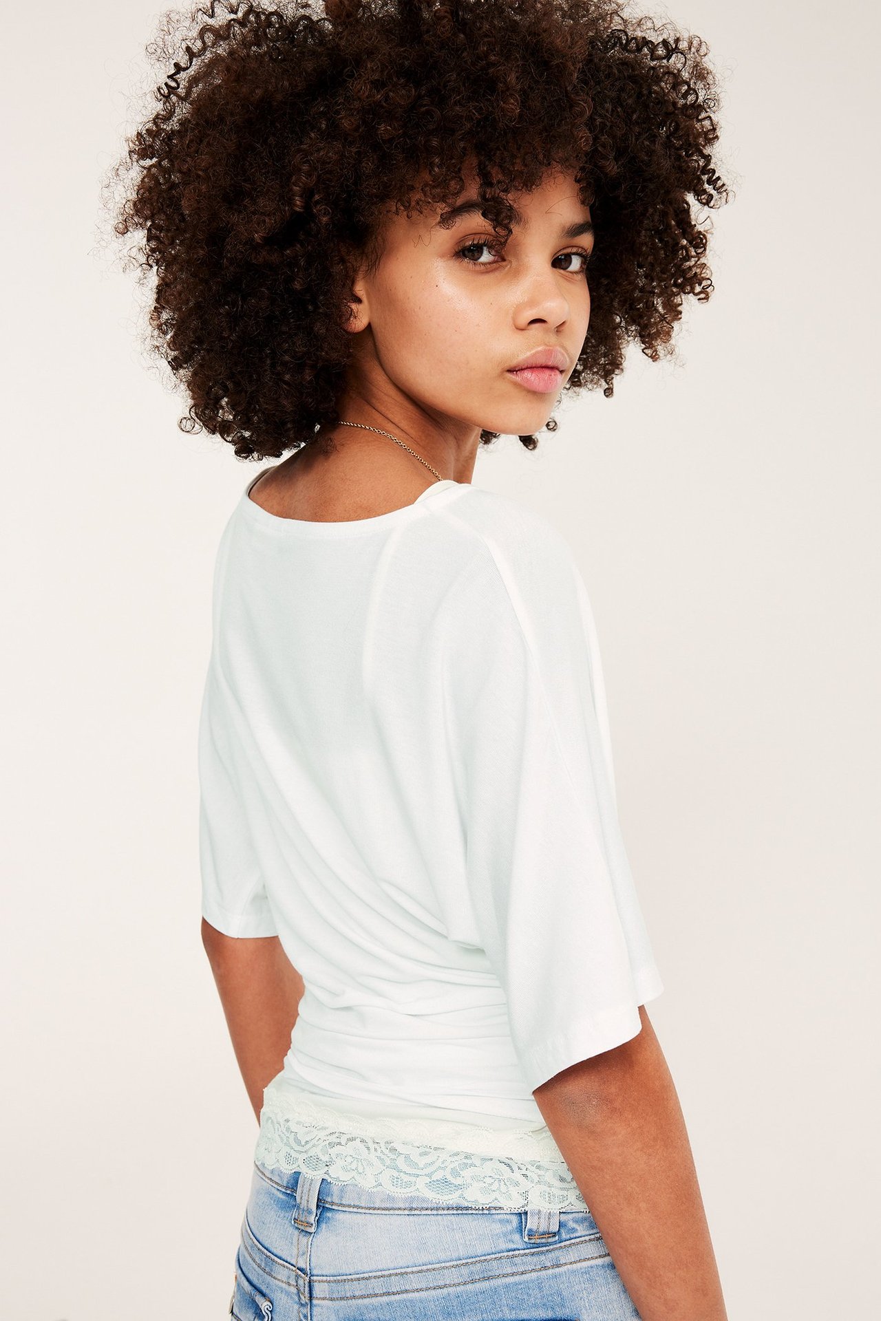 Kvinna från baksidan i vit draperad rynkad topp, ljusblå jeans och guldkedja.