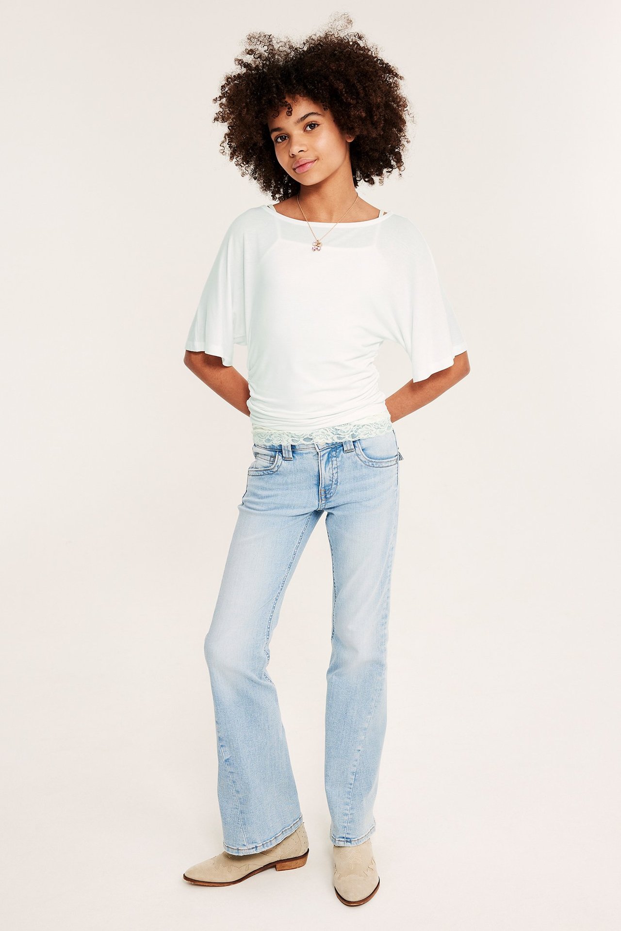 Framifrån: Ett barn bär en vit draperad topp med fjärilsärm, ljusblå utsvängda jeans och beige ankelboots.