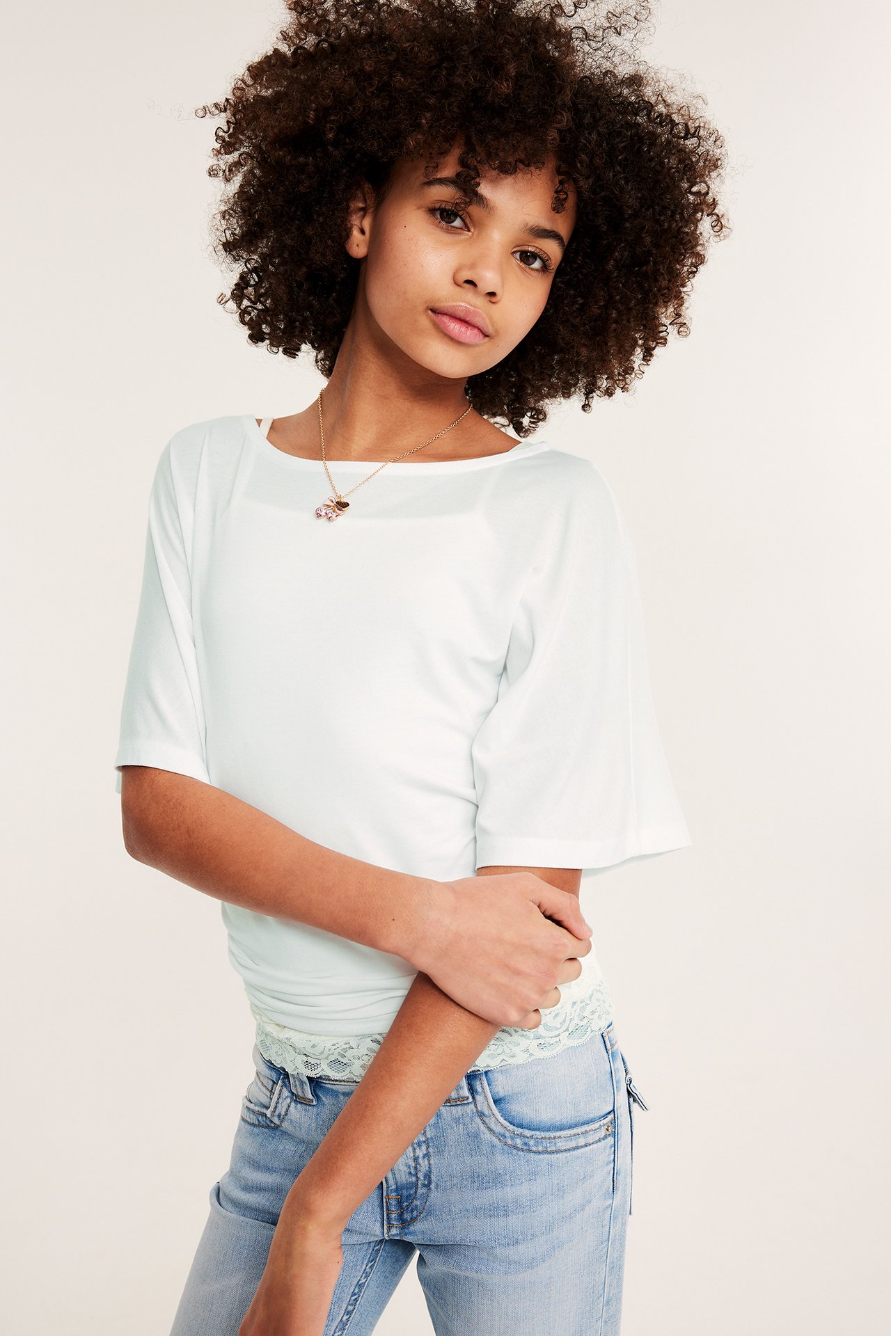 Framifrån: Ett barn bär en vit draperad topp med fjärilsärmar och rynkad midja, ljusblå jeans och ett guldhalsband.