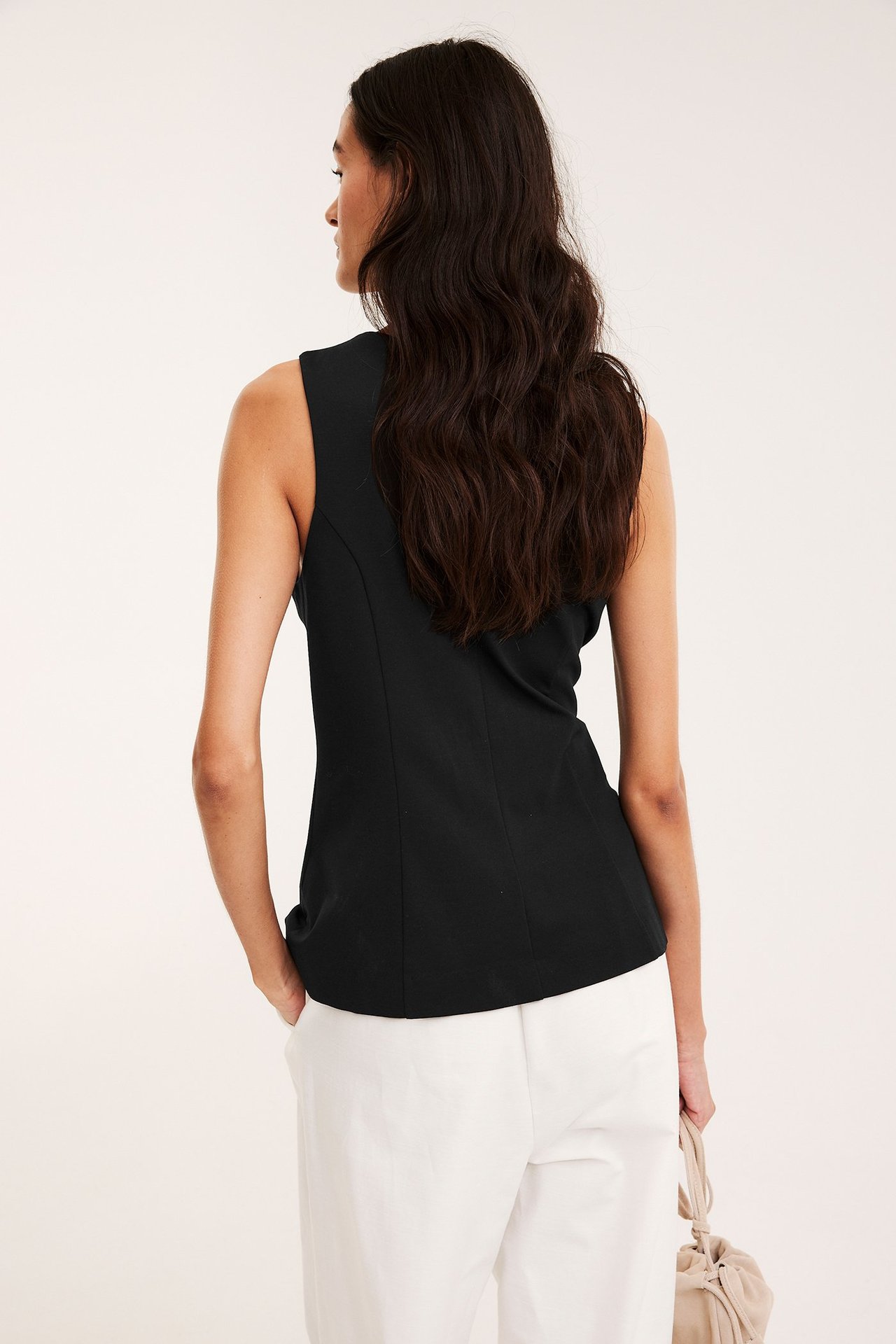 Ryggvy av kvinna i svart åtsittande väst, vita byxor, håller en beige handväska.