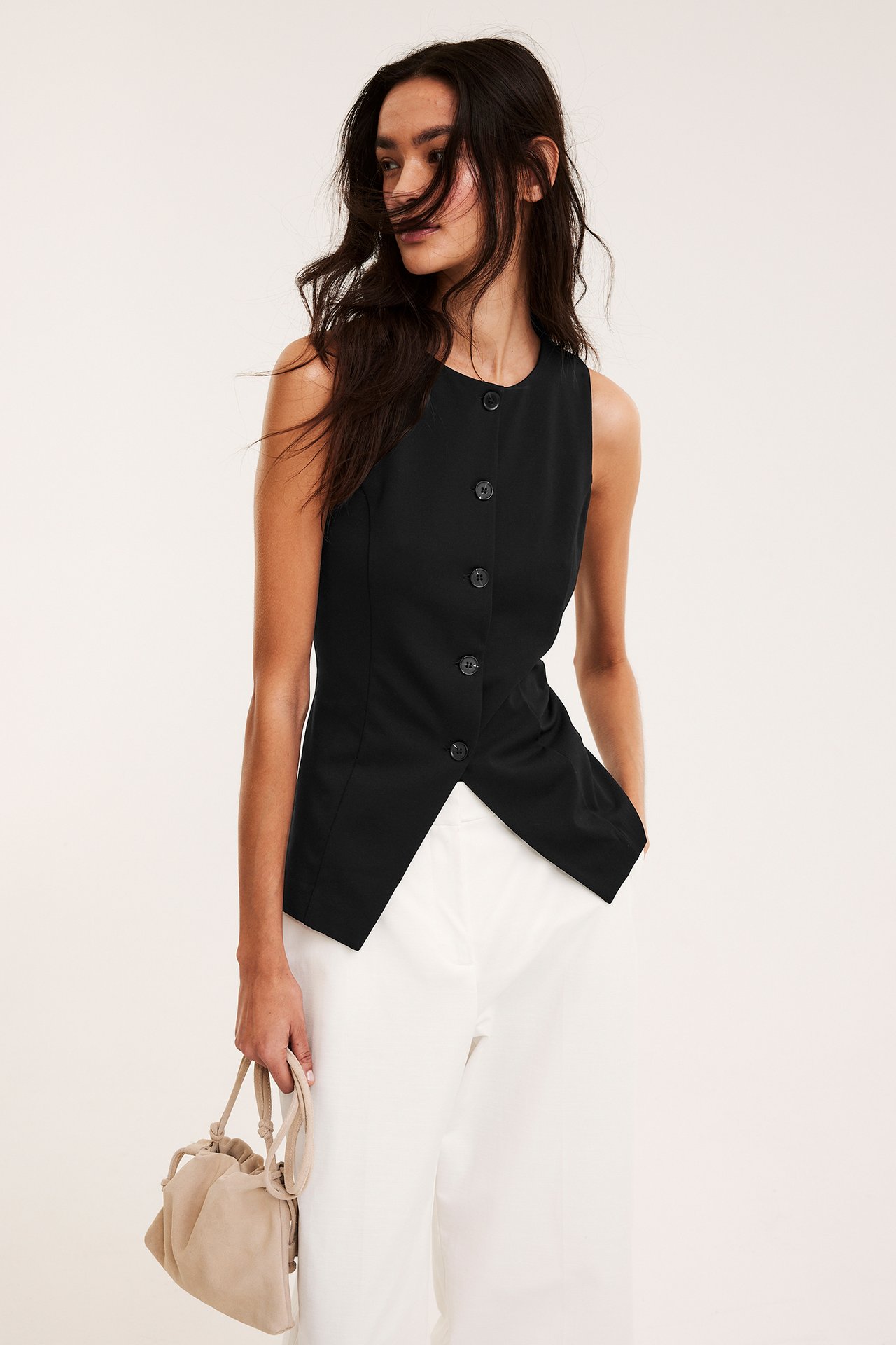 Edestä: Nainen istuvassa mustassa napitetussa liivissä, terävä helma, valkoiset leveät housut, pitää beigenväristä käsilaukkua.