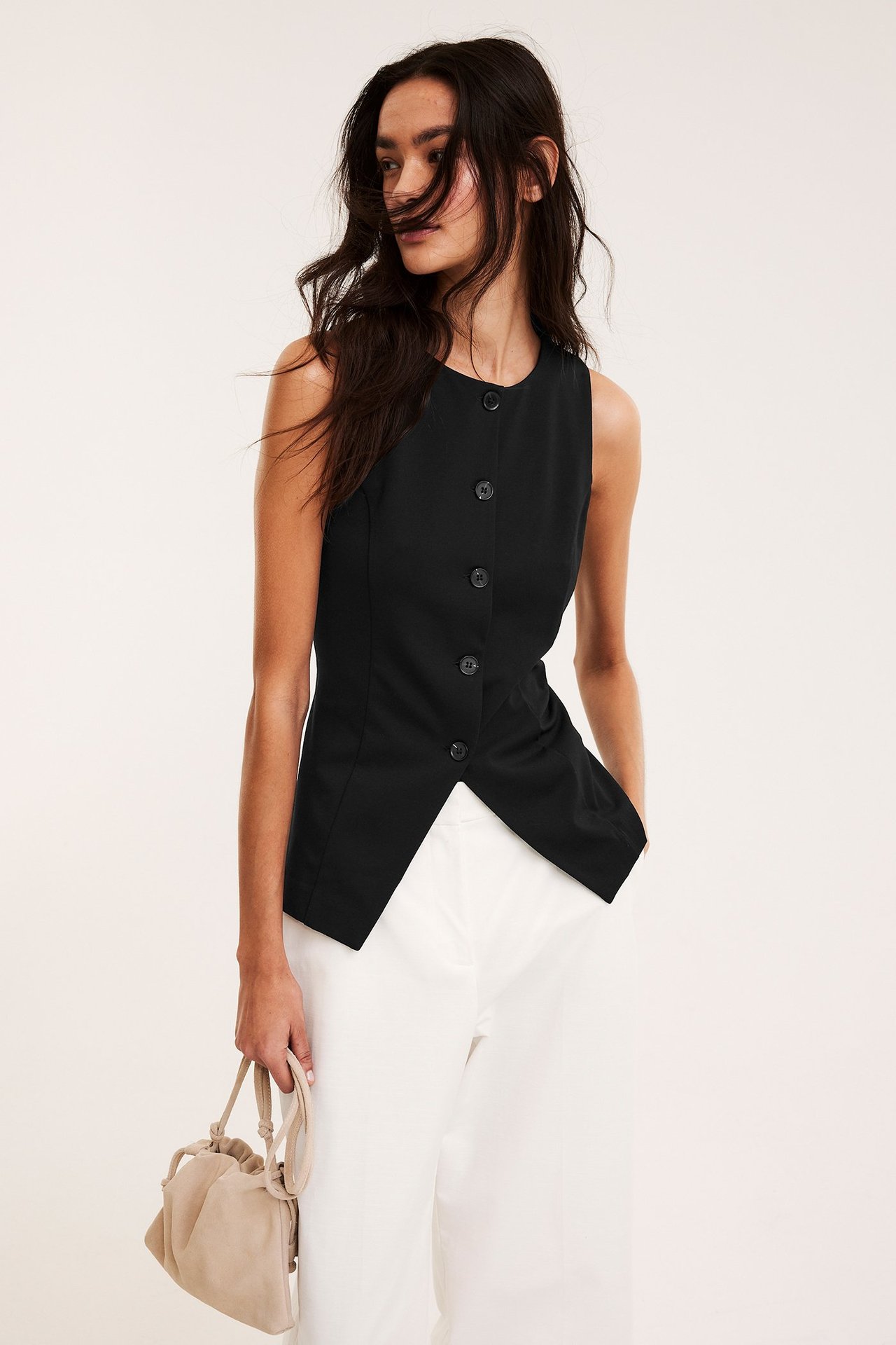 Widok z przodu: Kobieta w dopasowanej czarnej kamizelce, spiczasty dół, białych spodniach, trzyma beżową torebkę.