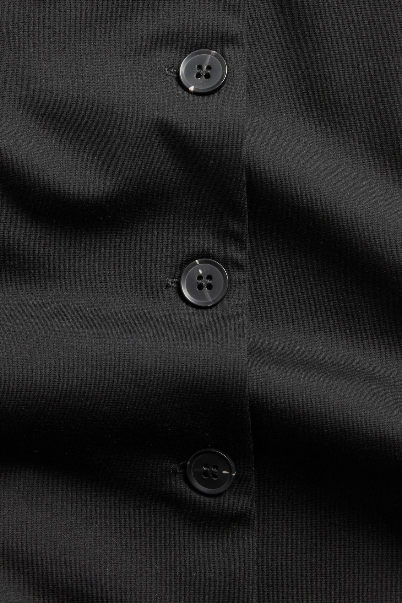 Lähikuva mustasta räätälöidystä liivistä, jossa kolme pyöreää, tummaa nappia nappilistassa.