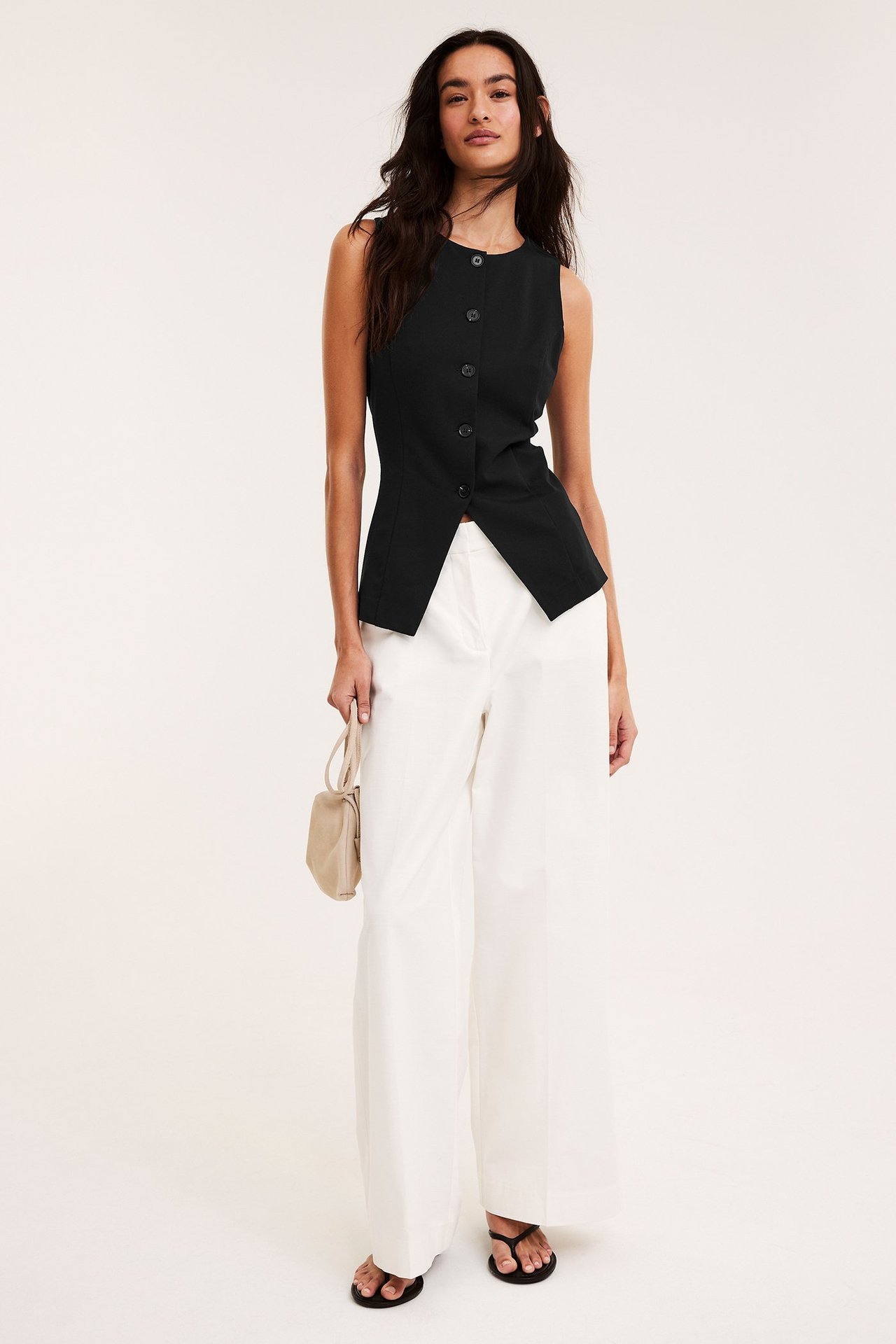 Widok z przodu: Kobieta w dopasowanej czarnej kamizelce, białych szerokich spodniach, czarnych klapkach, z beżową torebką.
