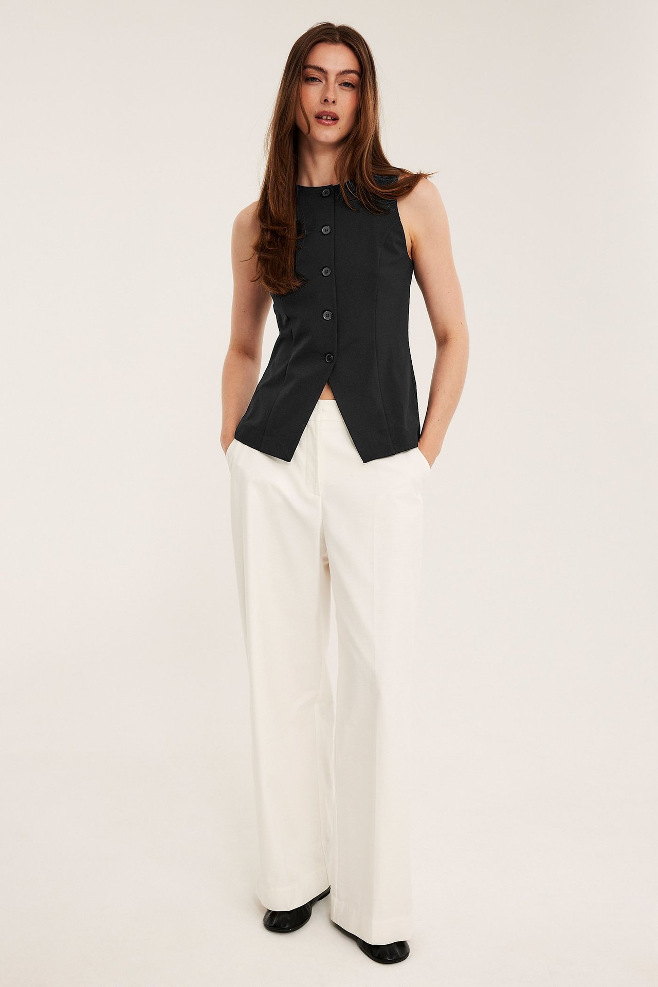 Widok z przodu kobiety w czarnej dopasowanej kamizelce na guziki, białych szerokich spodniach i czarnych butach.