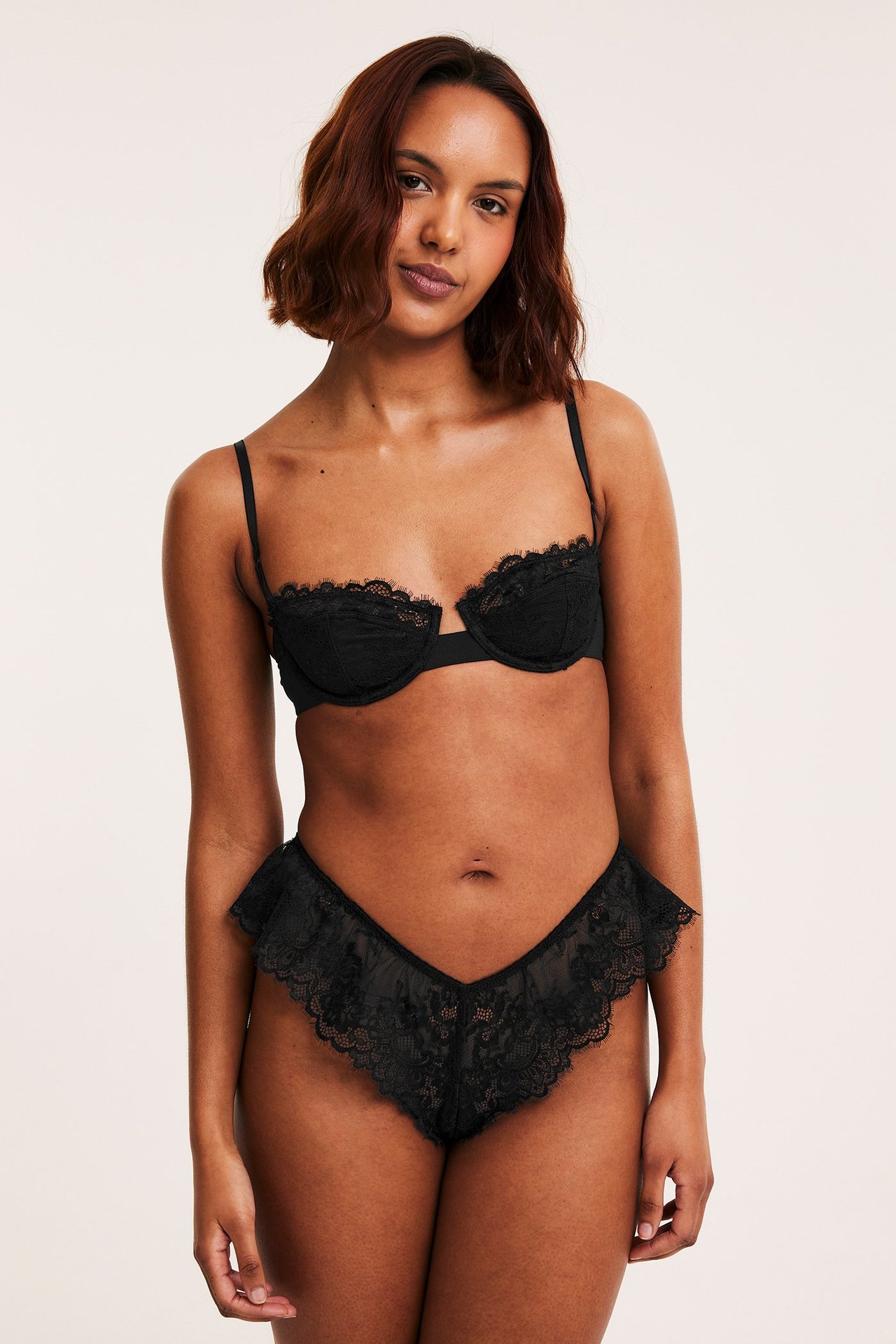 Etukuva naisesta, jolla on mustat pitsiset cheeky-alushousut röyhelöreunoilla ja yhteensopivat mustat pitsiliivit.