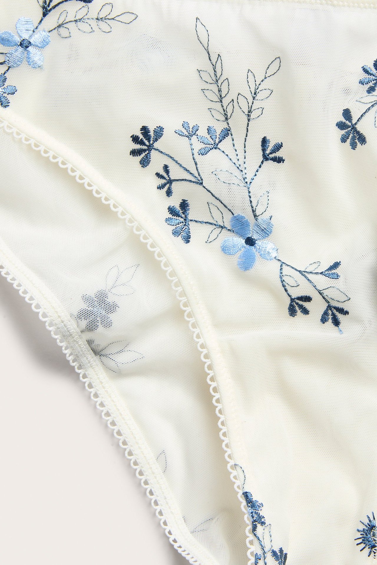 Nærbilde av offwhite mesh Brazilian-truse med blå blomsterbroderi og picot-elastisk kant.