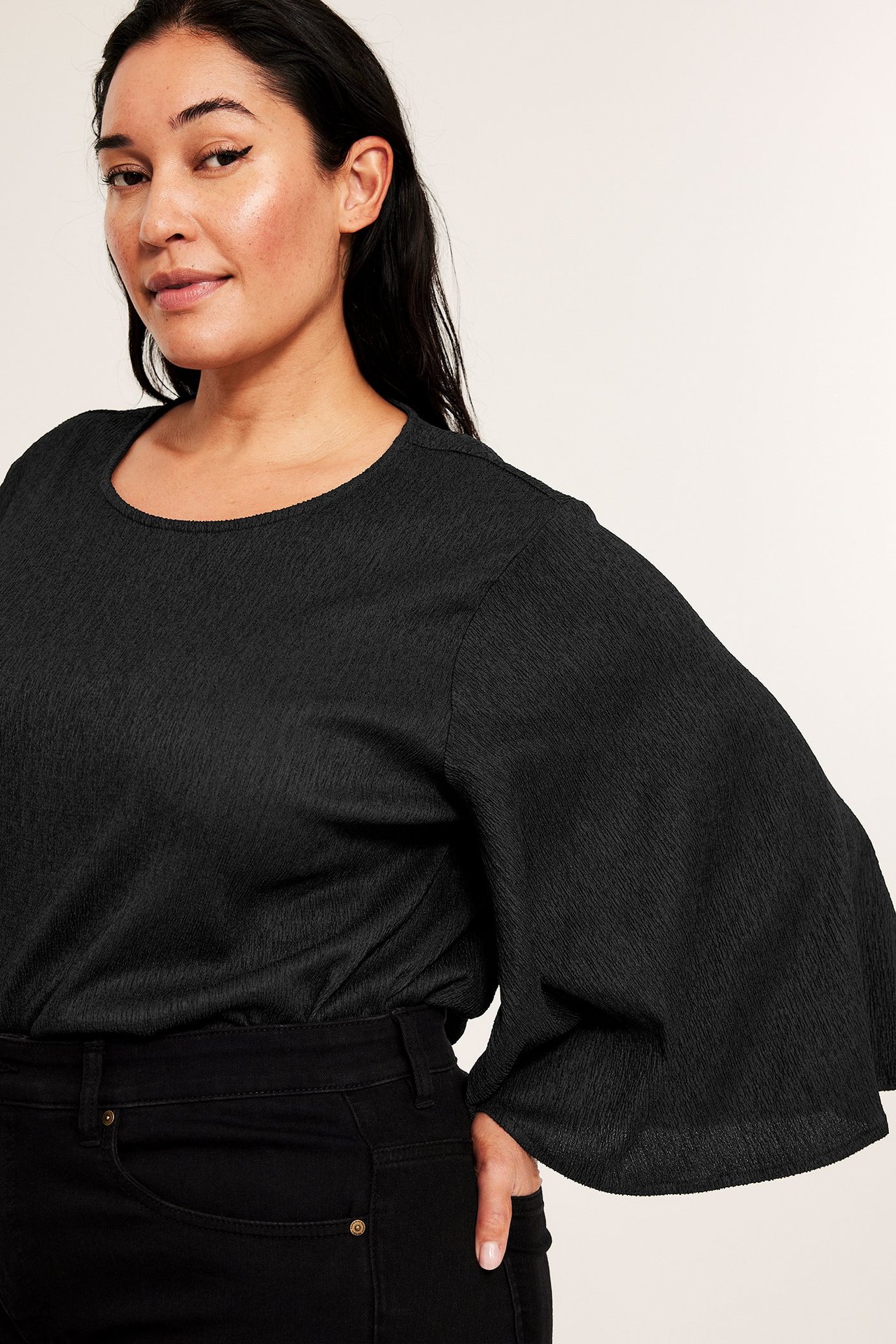 Framifrån: Kvinna i svart strukturerad festtopp med rund hals, långa fjärilsärmar, med svarta byxor.