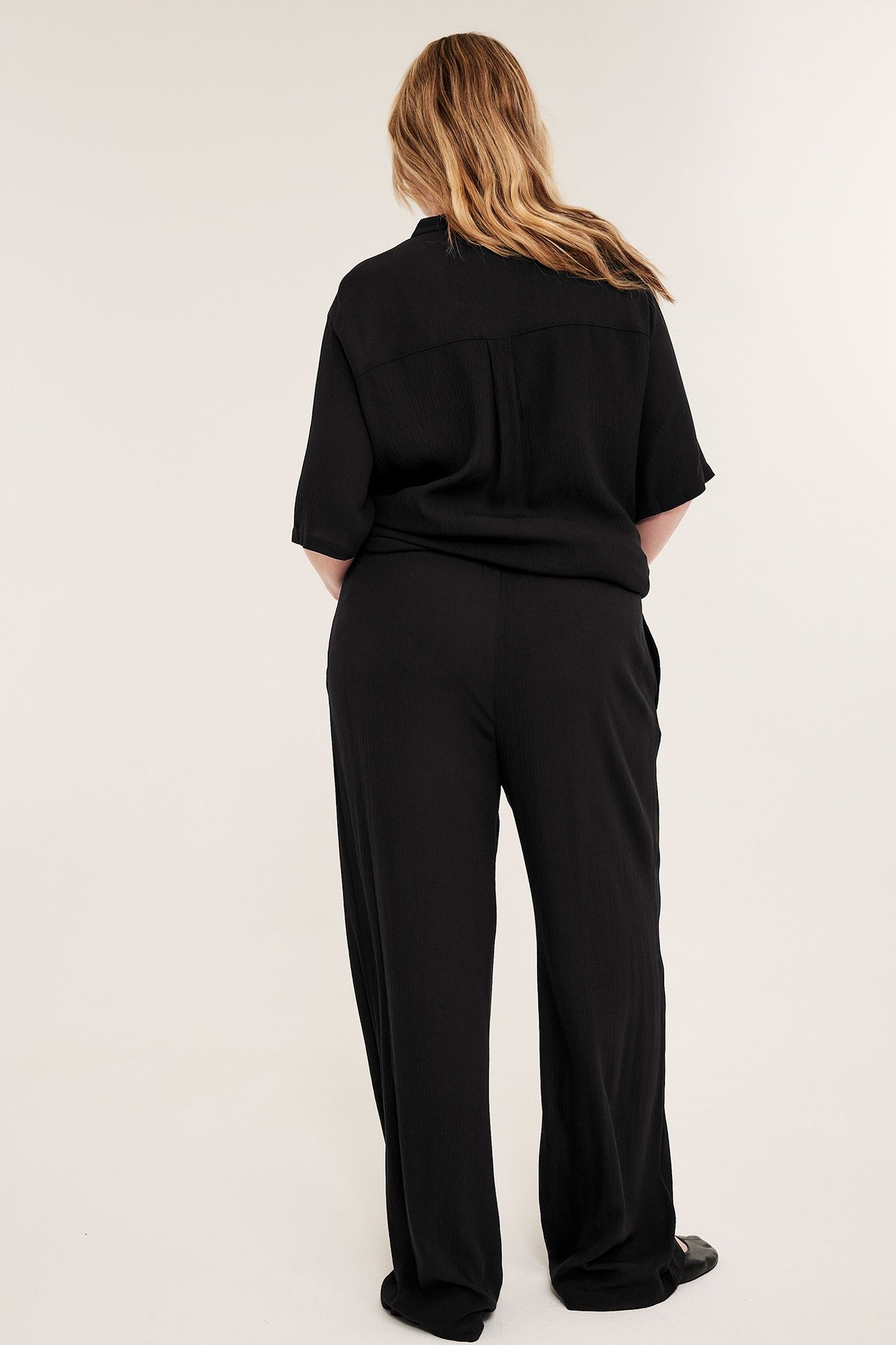 Bakfra: kvinne i svarte krøllete vide viskose pull-on-bukser og en matchende svart kortermet topp.