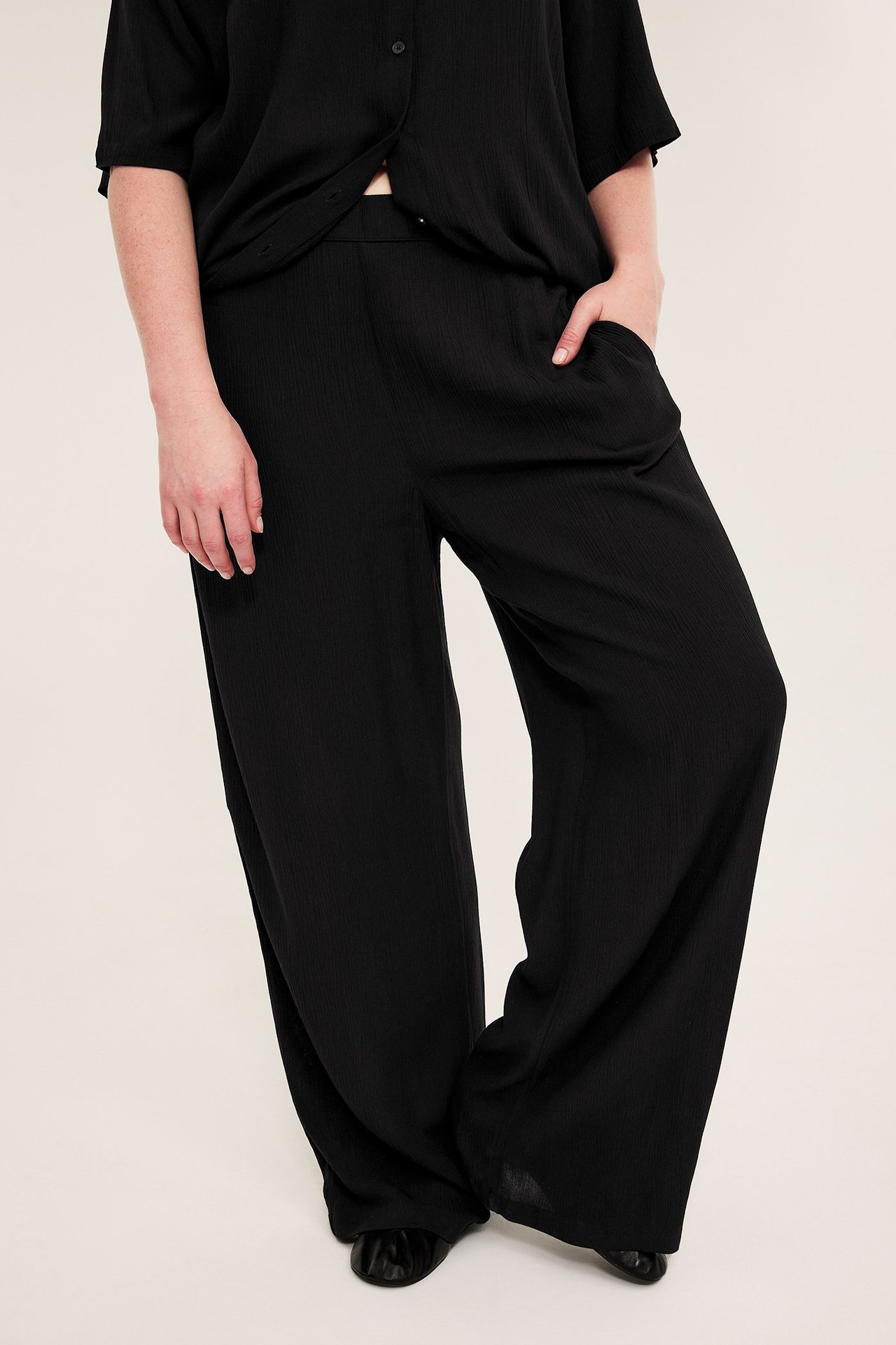 Framsida: kvinna i svarta krinklade vida byxor i viskos med sidofickor och en svart skjorta.