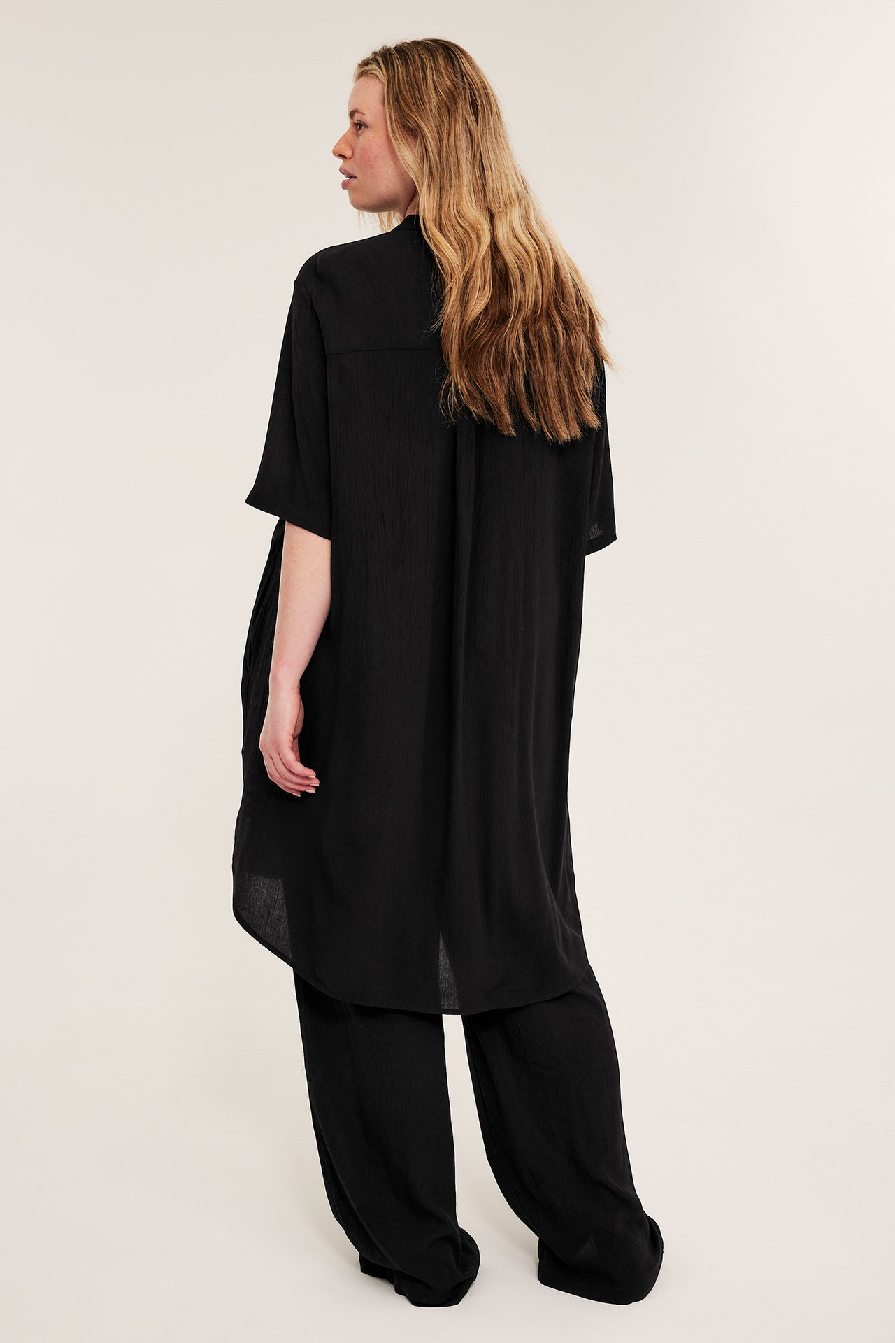 Kvinna bakifrån bär en svart kraglös lång lös viskosskjorta över svarta vida byxor.