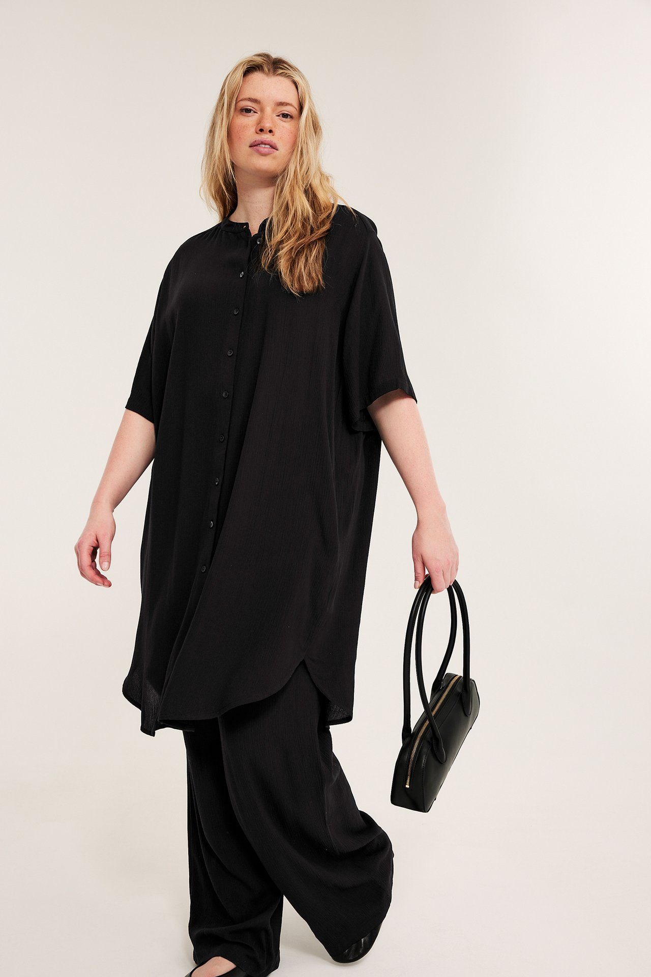 Frontvisning: kvinne i svart løs krageløs lang viskoseskjorte, svarte vide bukser, holder veske.