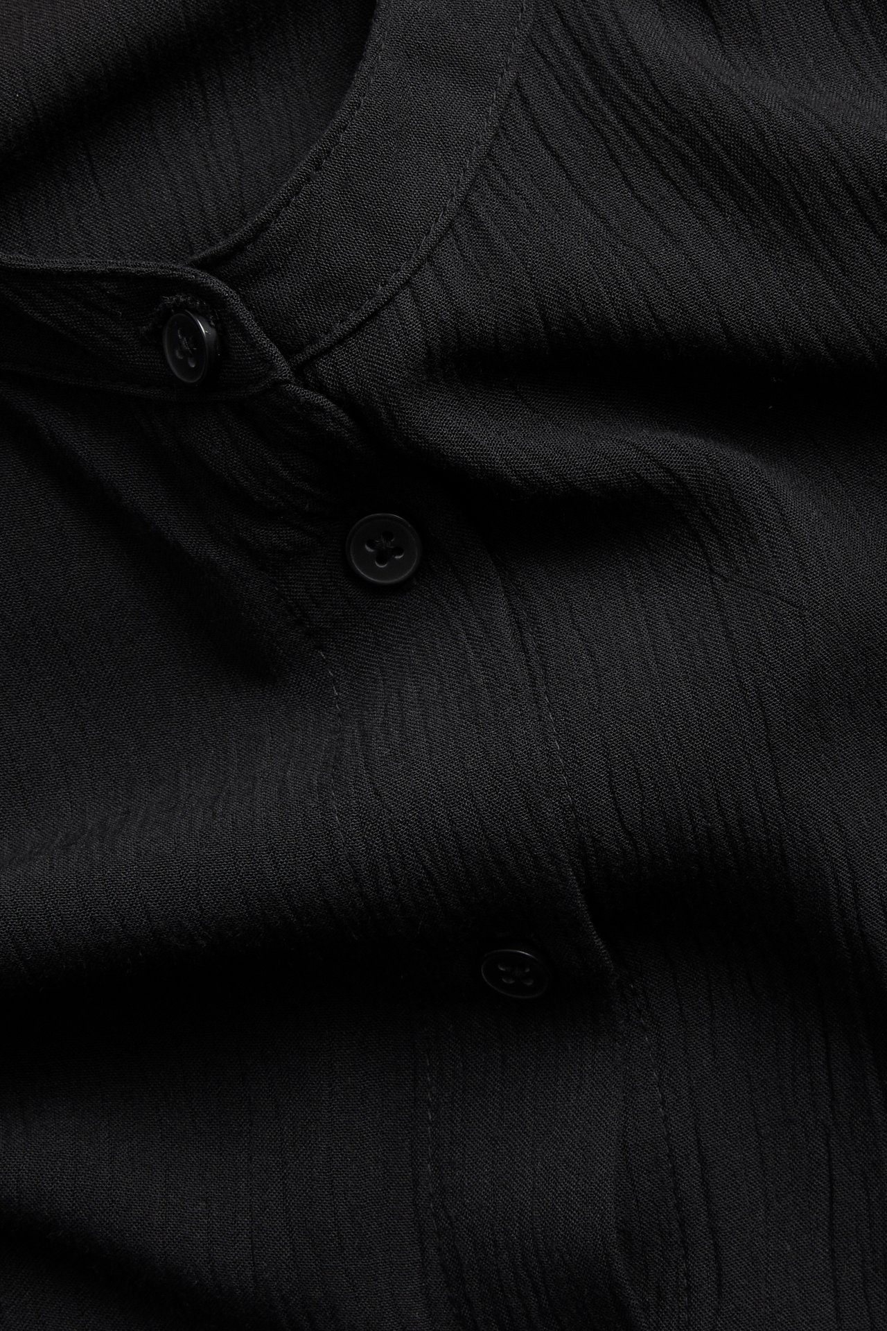 Närbild av en svart kraglös lös damskjorta i viskos med textur och synliga knappar.