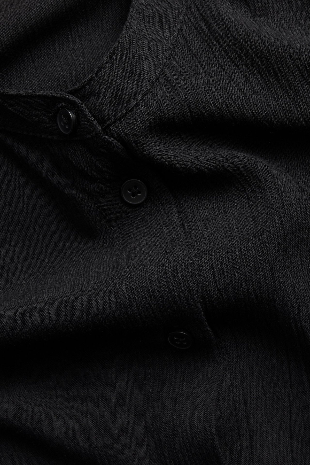 Zbliżenie czarnej, luźnej koszuli damskiej z wiskozy, bez kołnierza, z teksturowanej tkaniny, widoczne guziki.