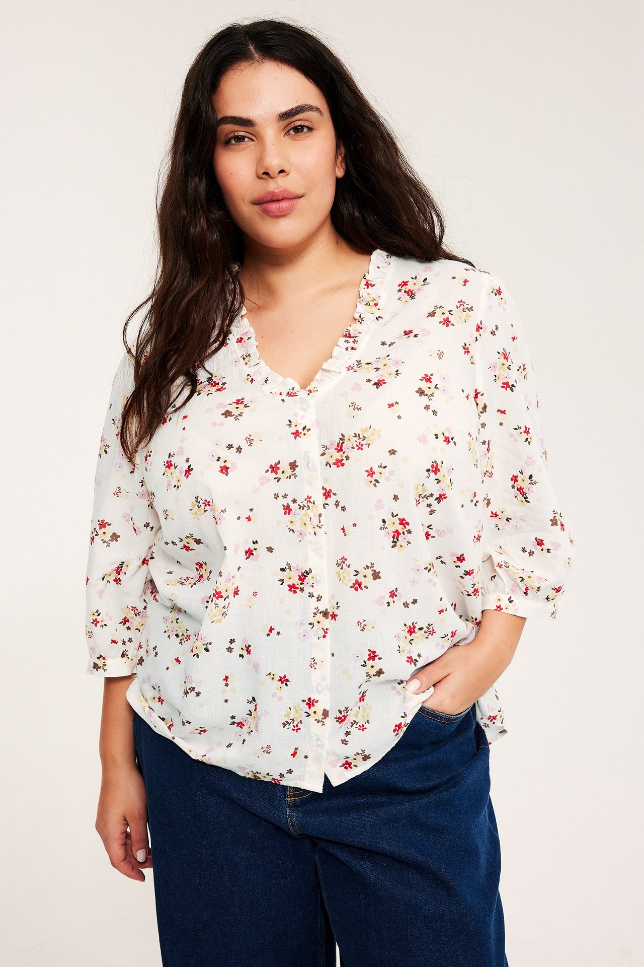 Nainen luonnonvalkoisessa kukkakuosisessa puuvillapuserossa, jossa röyhelö-V-pääntie, pussihihat ja tummansiniset farkut.