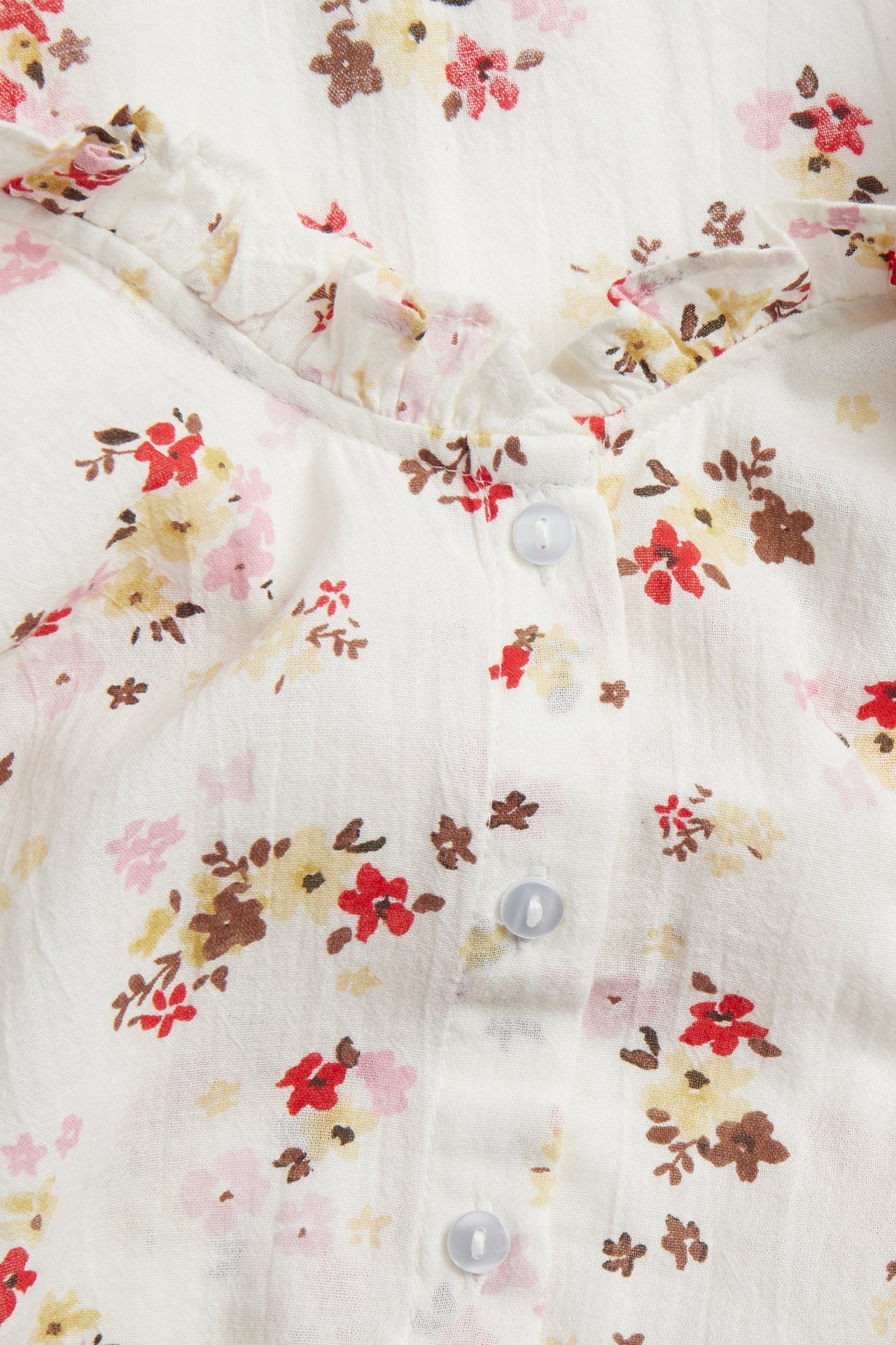 Lähikuva luonnonvalkoisesta puuvillapuserosta, jossa on röyhelö-V-pääntie, kukkakuvio ja vaaleansiniset napit.
