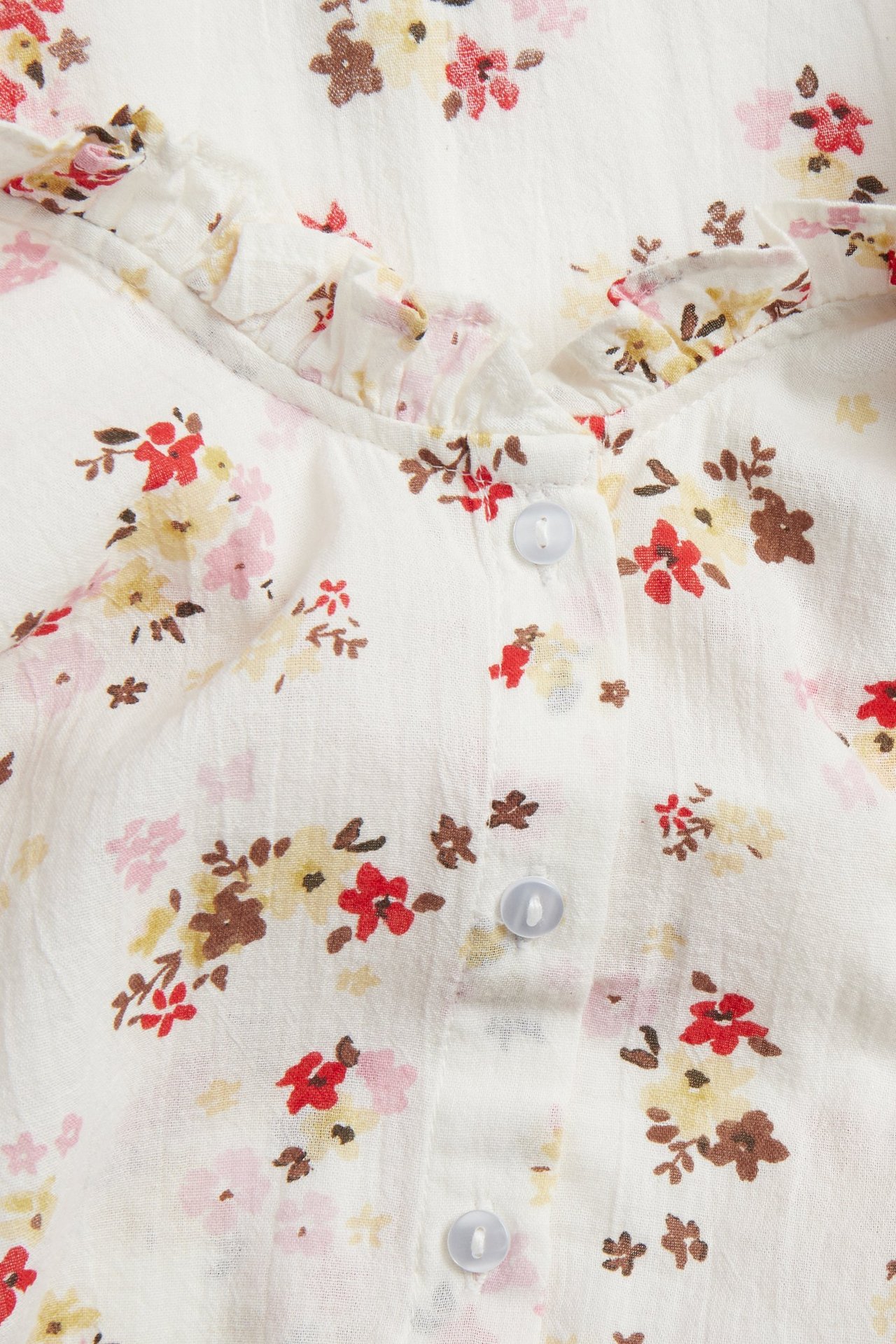 Zbliżenie na bawełnianą bluzkę w kolorze off-white z dekoltem V z falbanką, wzorem kwiatowym i jasnoniebieskimi guzikami.