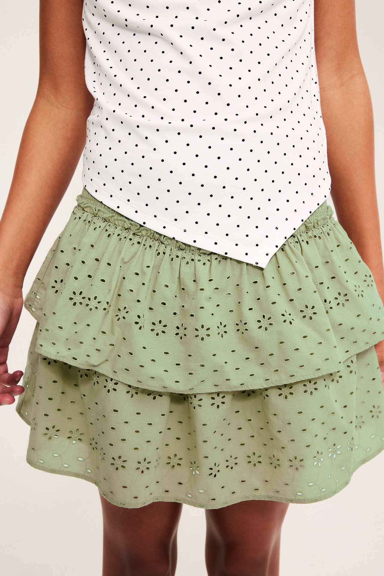 Forfra av et barn i mørkegrønt broderie anglaise-skjørt med volanger og hvit prikkete topp.