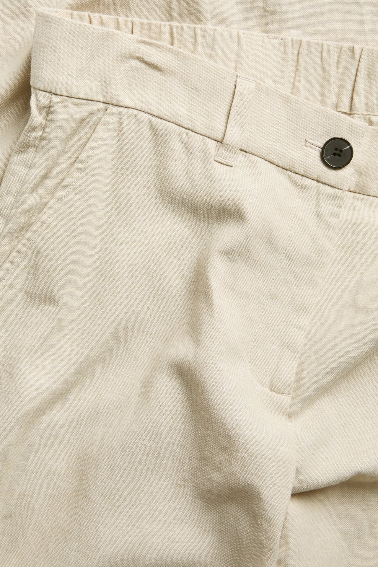 Lähikuva beigenvärisistä pellavasekoitteisista pukuhousuista, joissa joustava vyötärö takana ja tumma nappi.