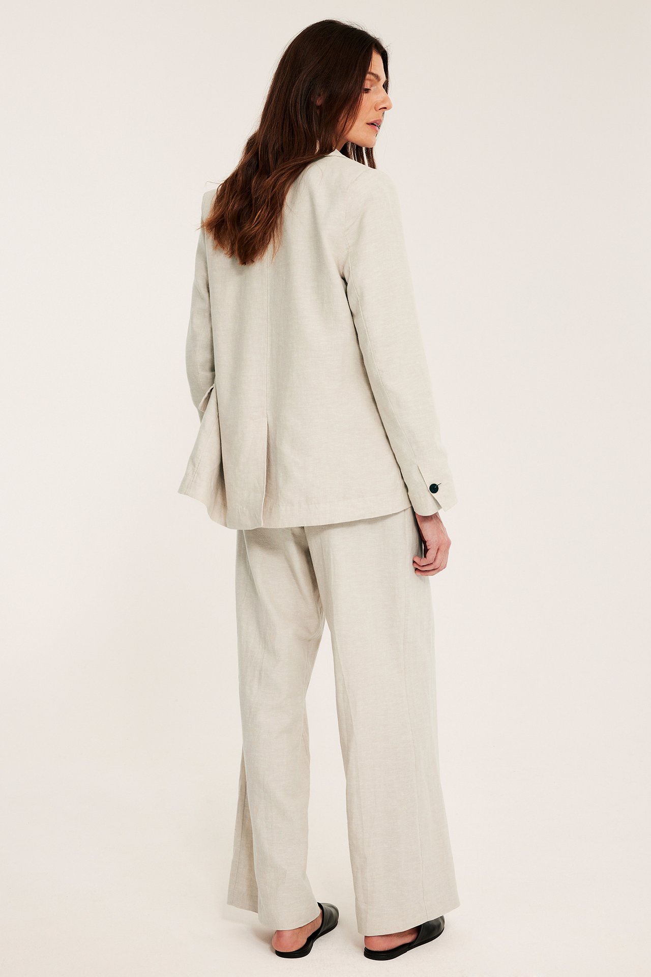 Bakfra av en kvinne i løs beige linblanding blazer, matchende bukser og svarte flate sko.