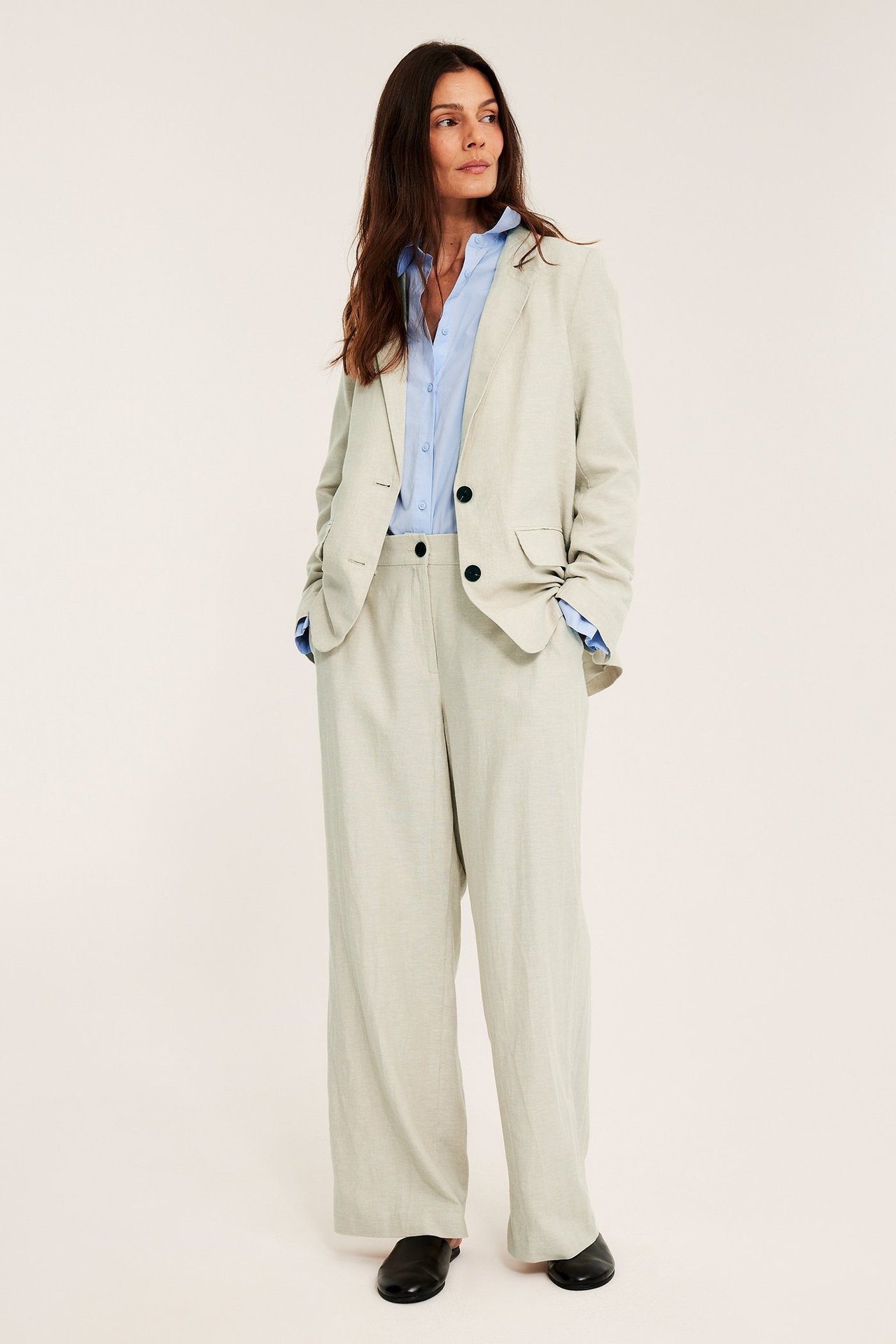 Frontutsikt: kvinne i lys beige melert linblanding blazer med mørke knapper, blå skjorte, matchende bukser, sorte mules.
