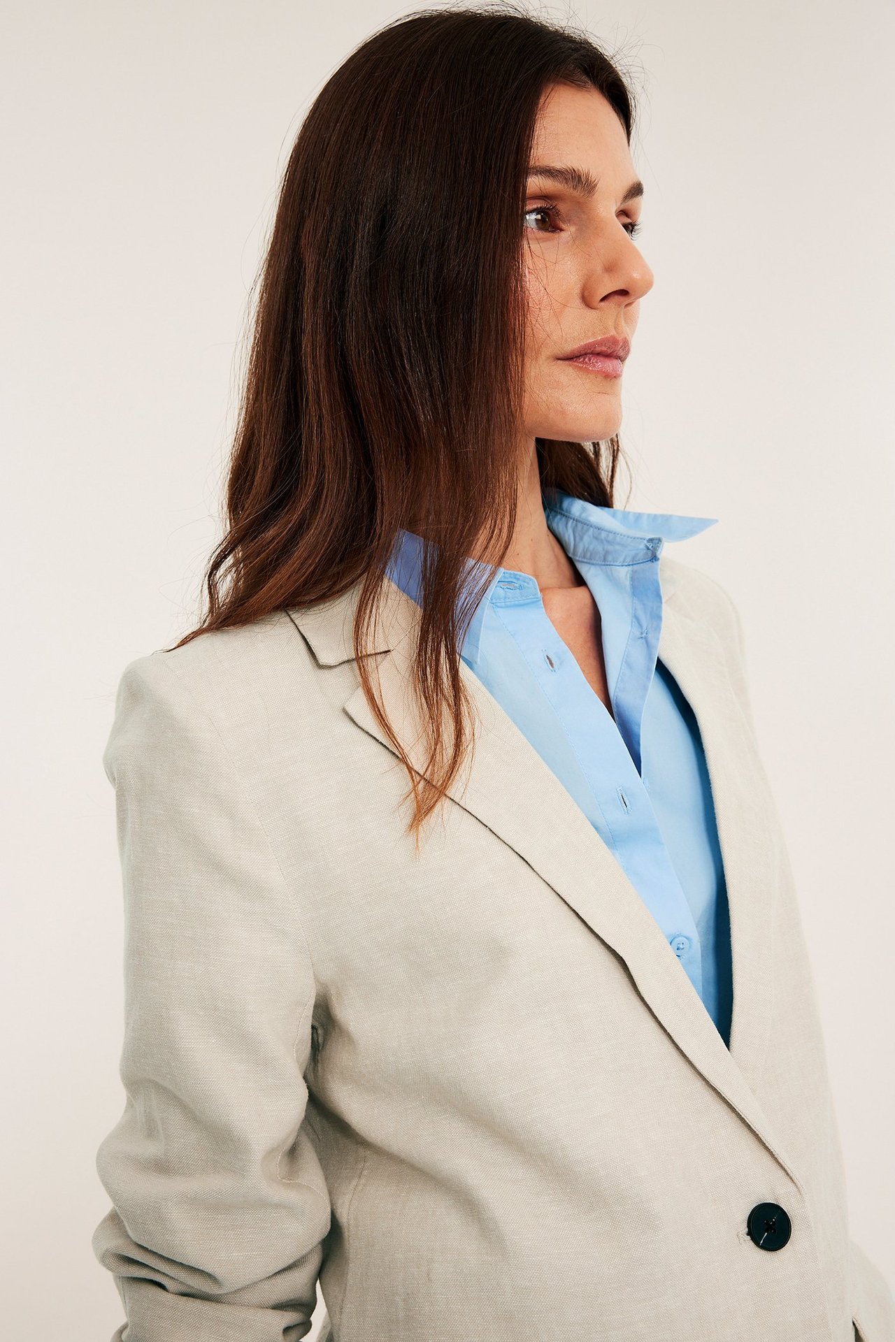 Widok z boku kobiety w otwartej beżowej melanżowej lnianej marynarce na jasnoniebieskiej koszuli.